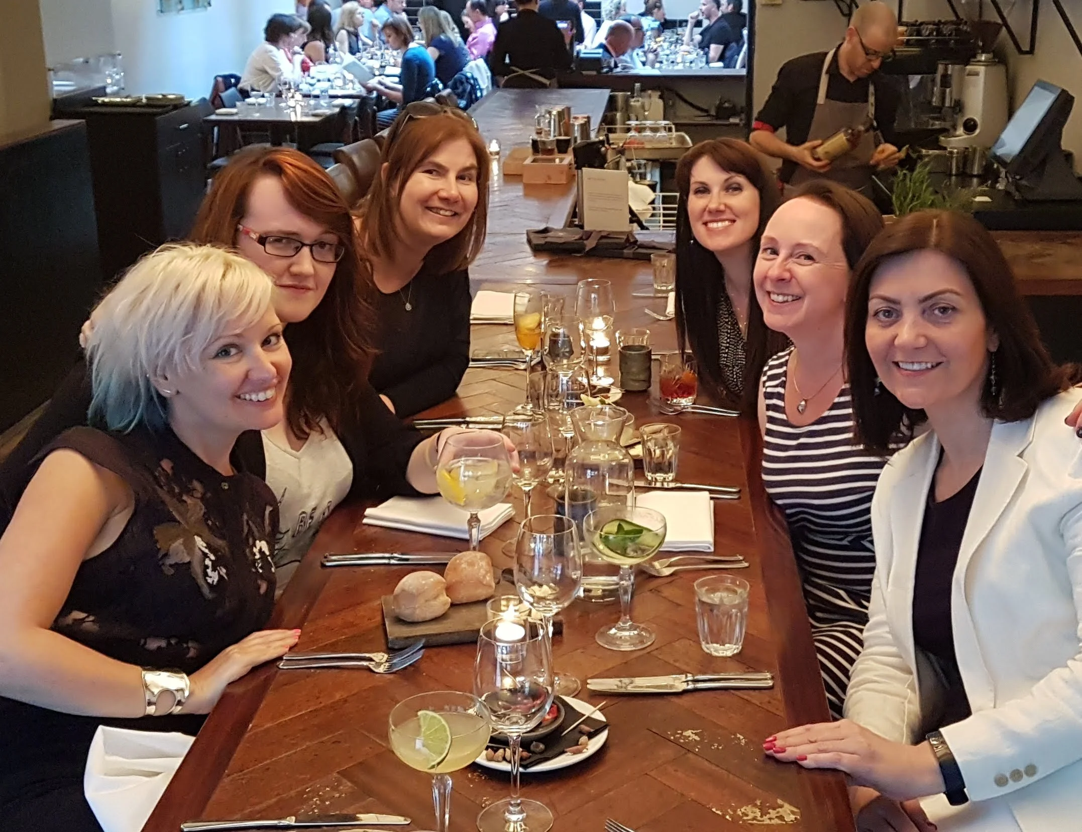Six women seated at a wooden dining table in a restaurant, smiling at the camera, with drinks and bread rolls in front of them. There are more people dining in the background.
