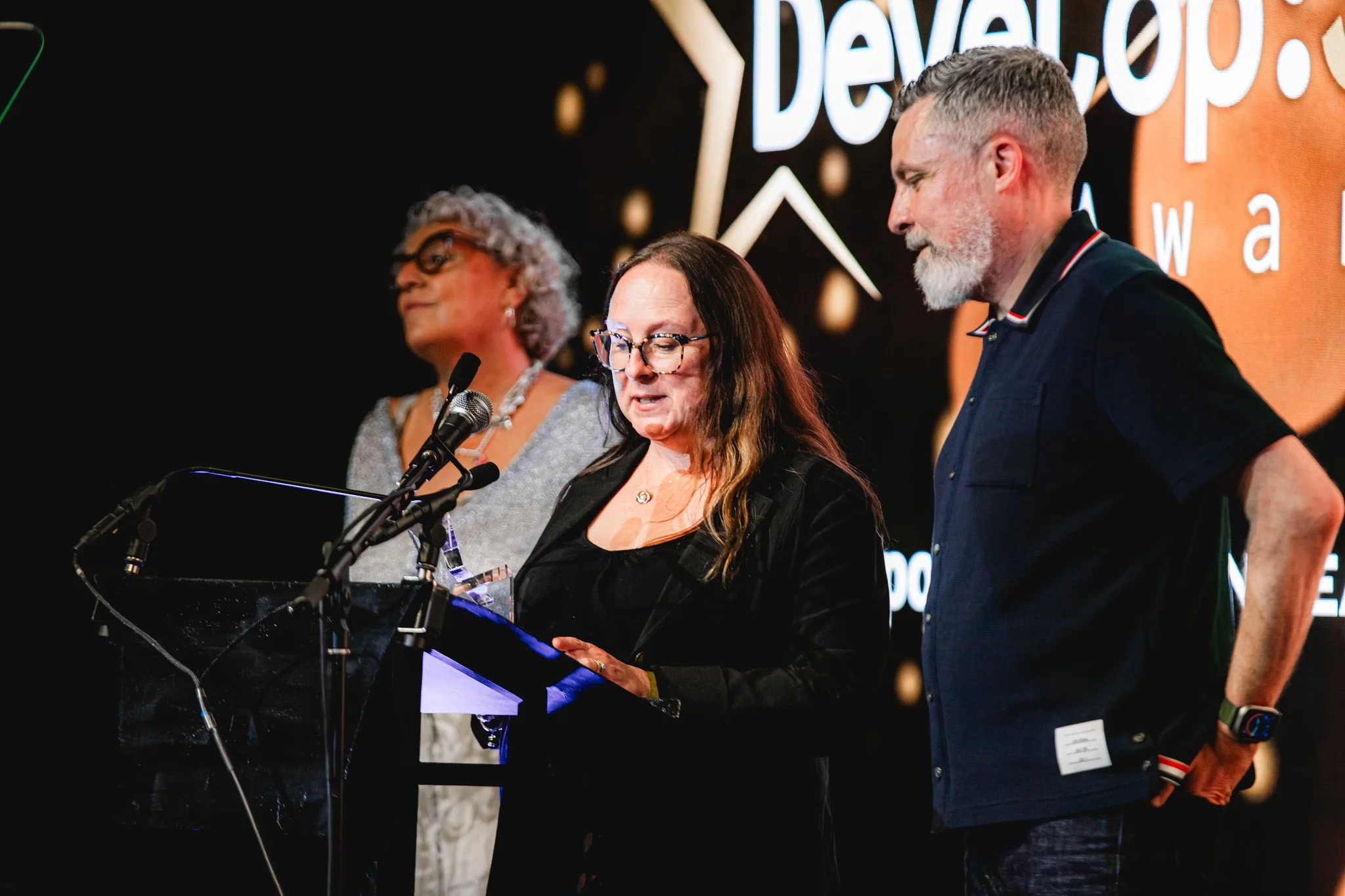 Group of four people standing on a stage at an event. The woman in the center, with long brown hair and glasses, is reading from a paper. To her right, a man with a gray beard and short hair is looking at her. To her left, a woman with curly gray hair and glasses is standing with her eyes closed. The background features a logo or sign with the word 'Development' visible.