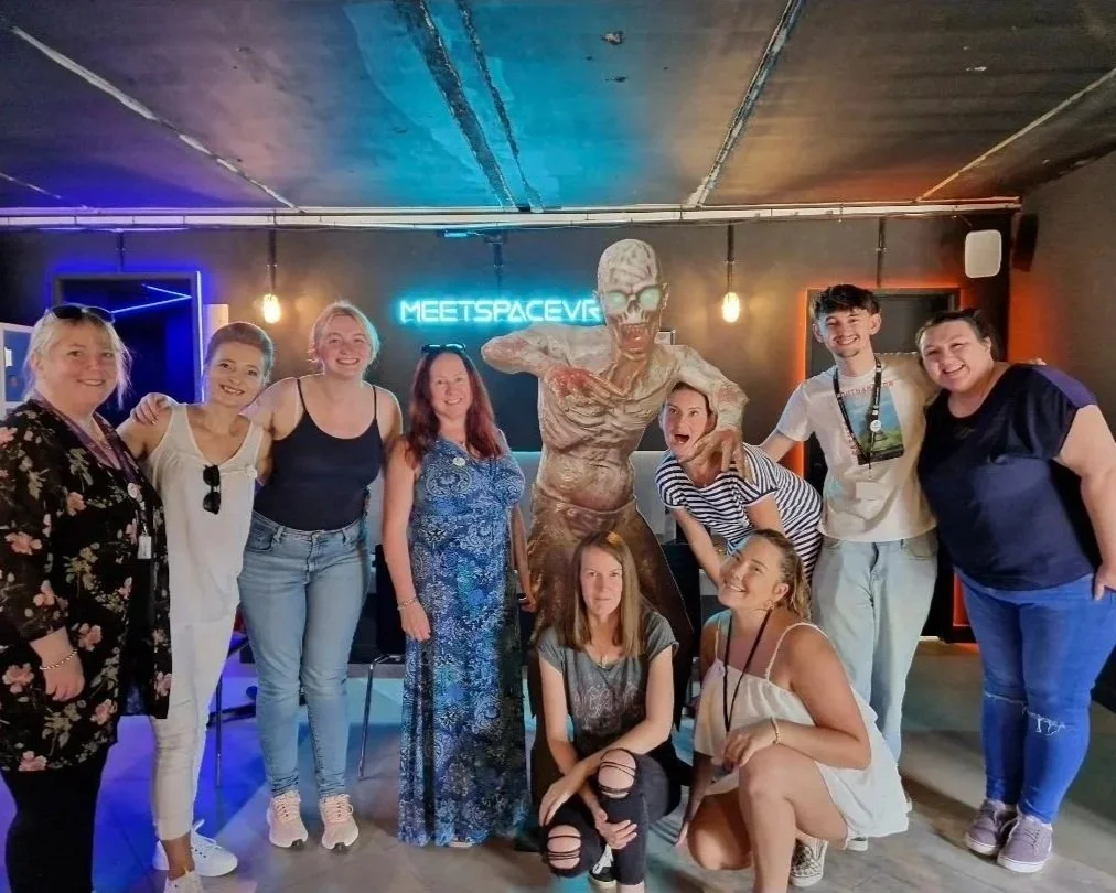 Group of people posing next to a zombie sculpture with a neon sign that says 'MEETSPACEVR' in a dark, modern room with industrial decor.