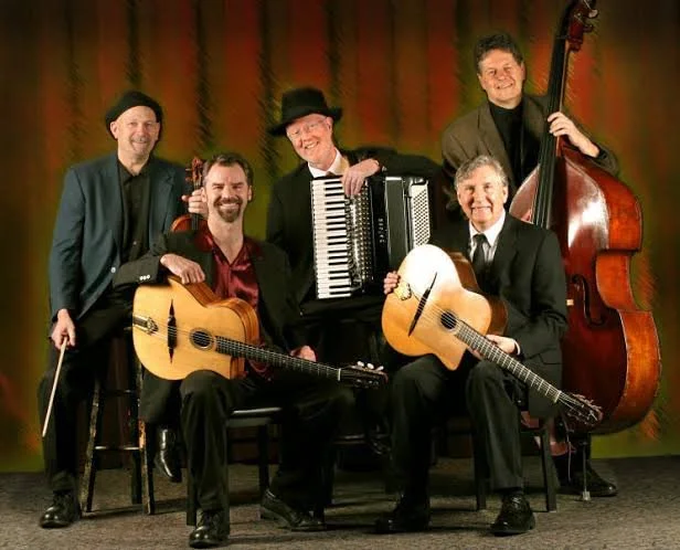 Five men in formal attire holding musical instruments, including a violin, acoustic guitars, accordion, and upright bass, posed in front of a colorful curtain.