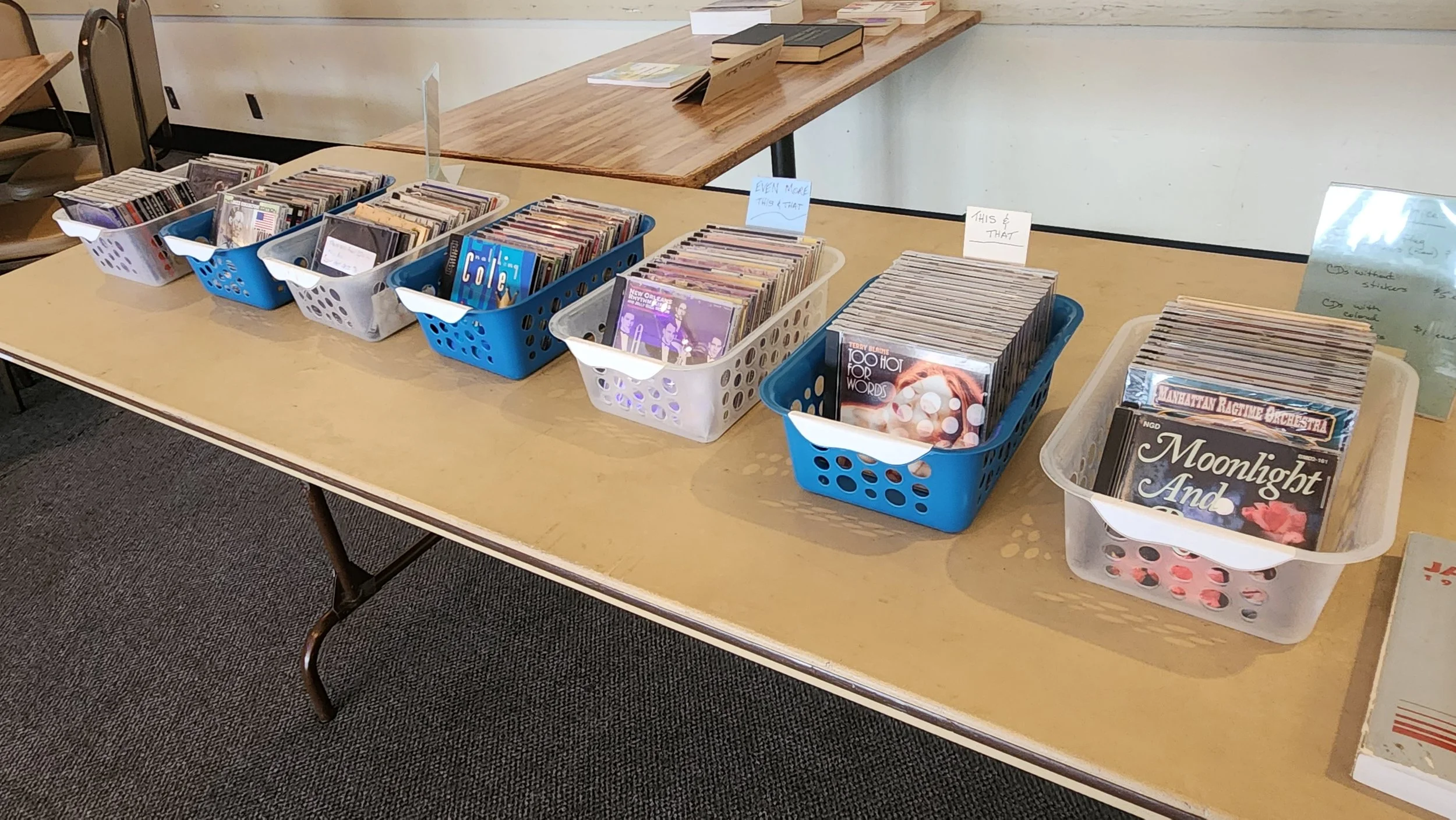 Table with several baskets of used books for sale, organized in rows, in an indoor setting.