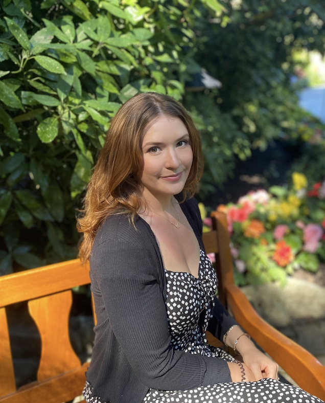 A young woman with auburn hair sitting on a wooden bench outdoors, surrounded by greenery and colorful flowers.