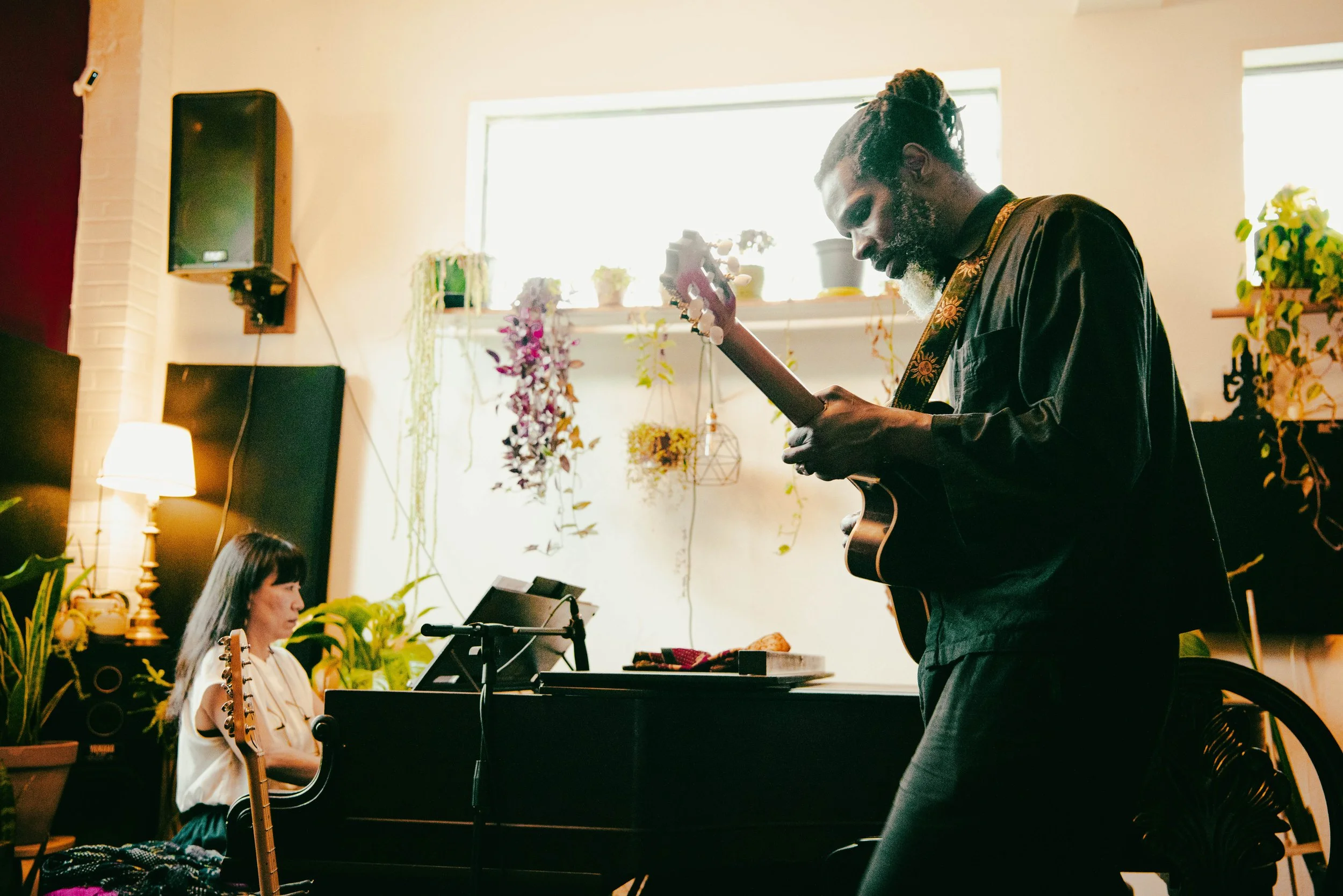 LuFuki playing an acoustic guitar and singing, and Aya Sekine playing a piano, performing indoors in a cozy, decorated room with plants and hanging flowers.