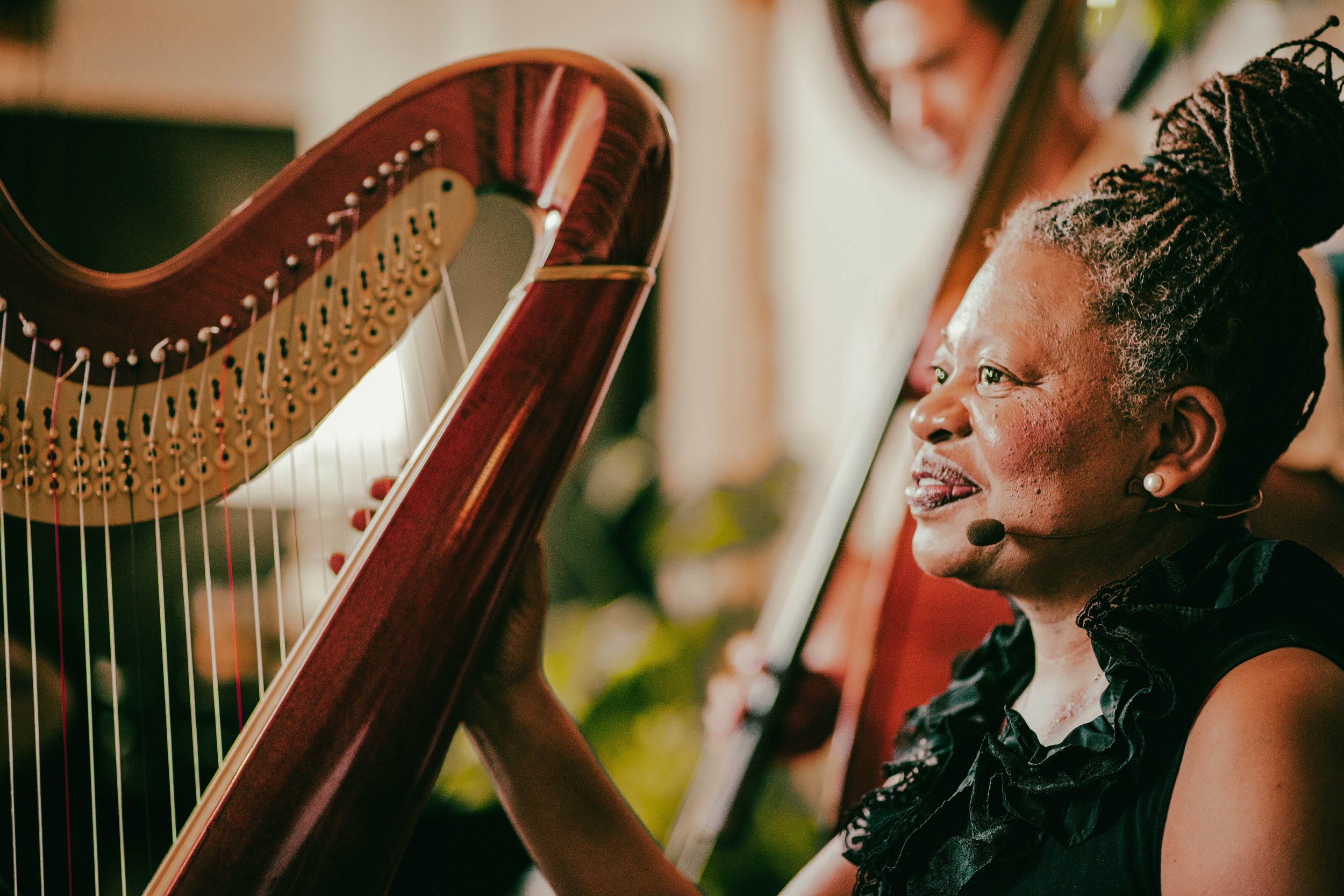 A woman playing a harp with a microphone headset on, smiling and looking at the harp, with another musician blurred in the background.