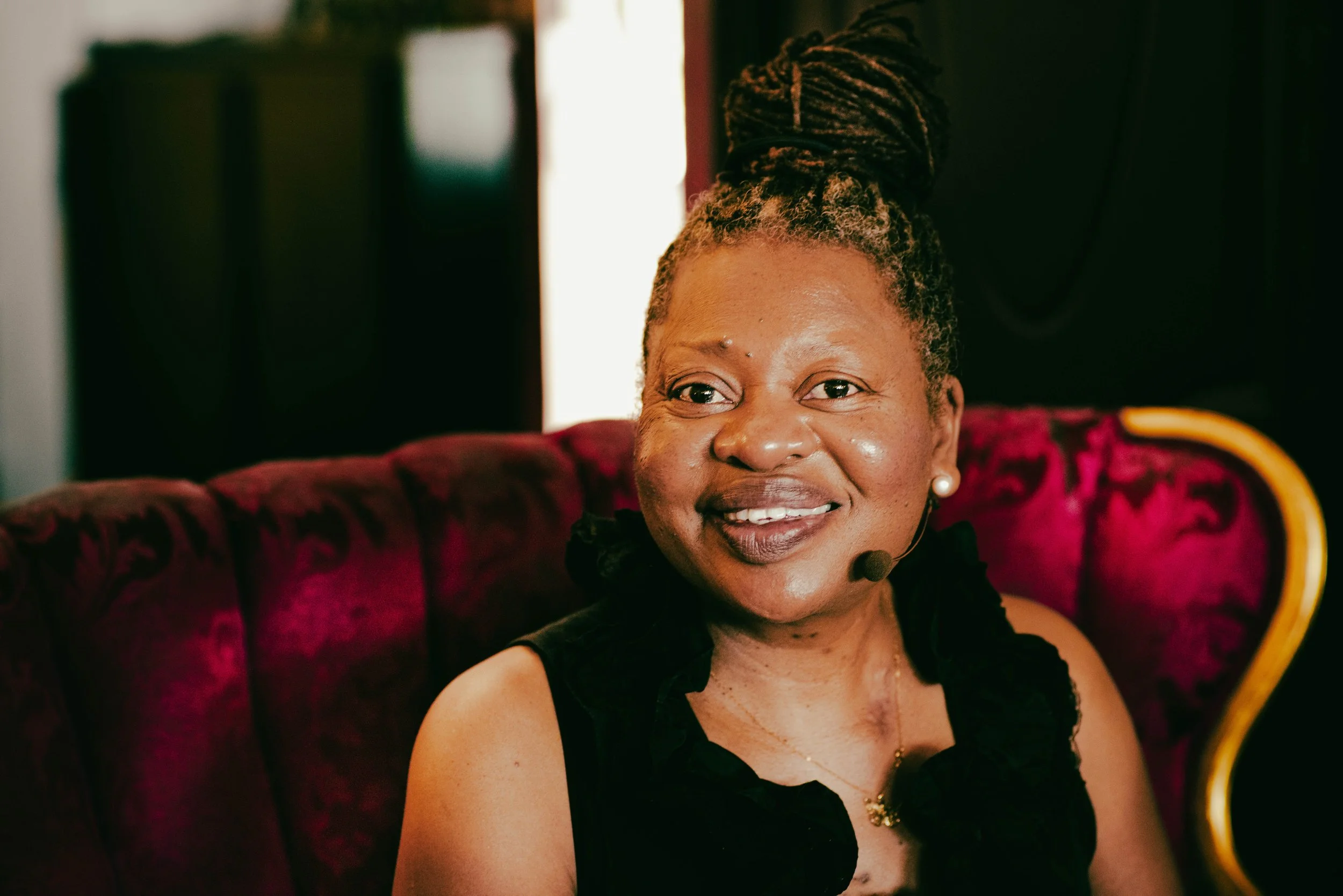Destiny Muhammad with hair tied up in a bun, wearing pearl earrings, a black top with ruffled shoulders, and a small microphone on her collar, sitting on a red velvet chair with gold trim.