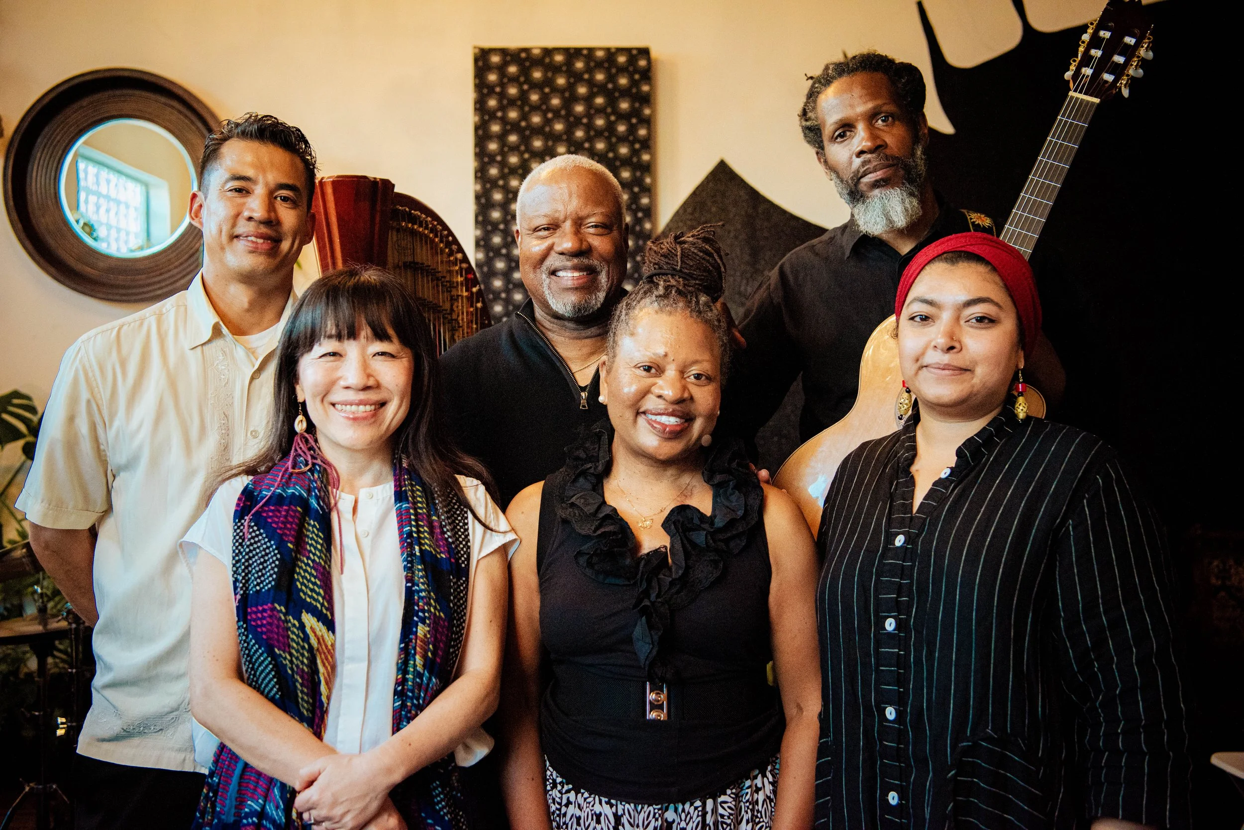 Group of six diverse musicians posing together indoors, with musical instruments including a guitar and harp in the background.