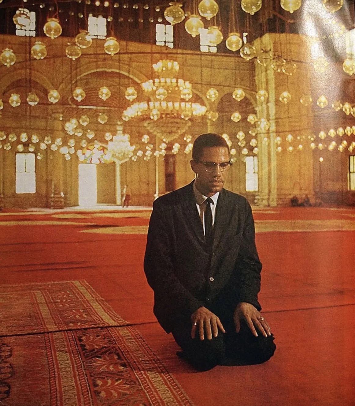 Malcolm X dressed in a black suit and glasses praying on a red carpet inside the Muhammad Ali mosque in Cairo, Egypt with chandeliers and ornate lighting fixtures.