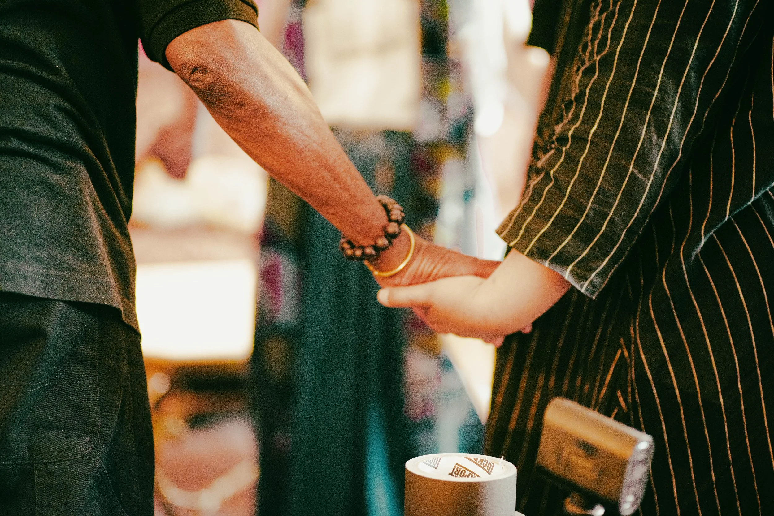 Two people holding hands in a gesture of connection or support, one wearing black with a beaded bracelet and a yellow thread bracelet, the other wearing a black pinstriped outfit, with a roll of brown paper tape on a wooden surface nearby.