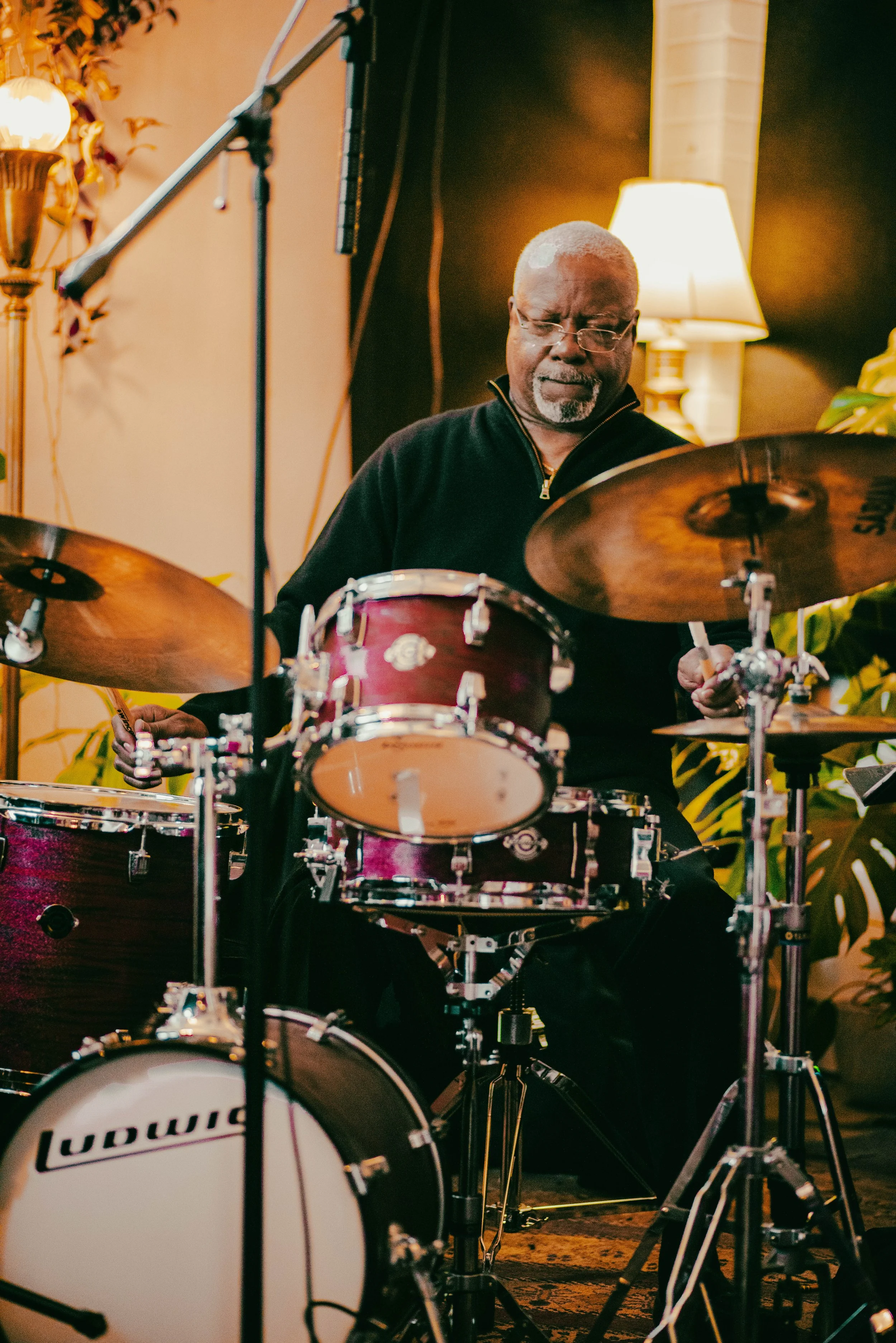 An elderly man with glasses and a white beard playing a drum set in a warmly lit room.