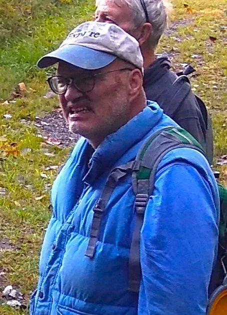 Two men outdoors in a forested area, wearing outdoor gear including backpacks and hats, with one man smiling.