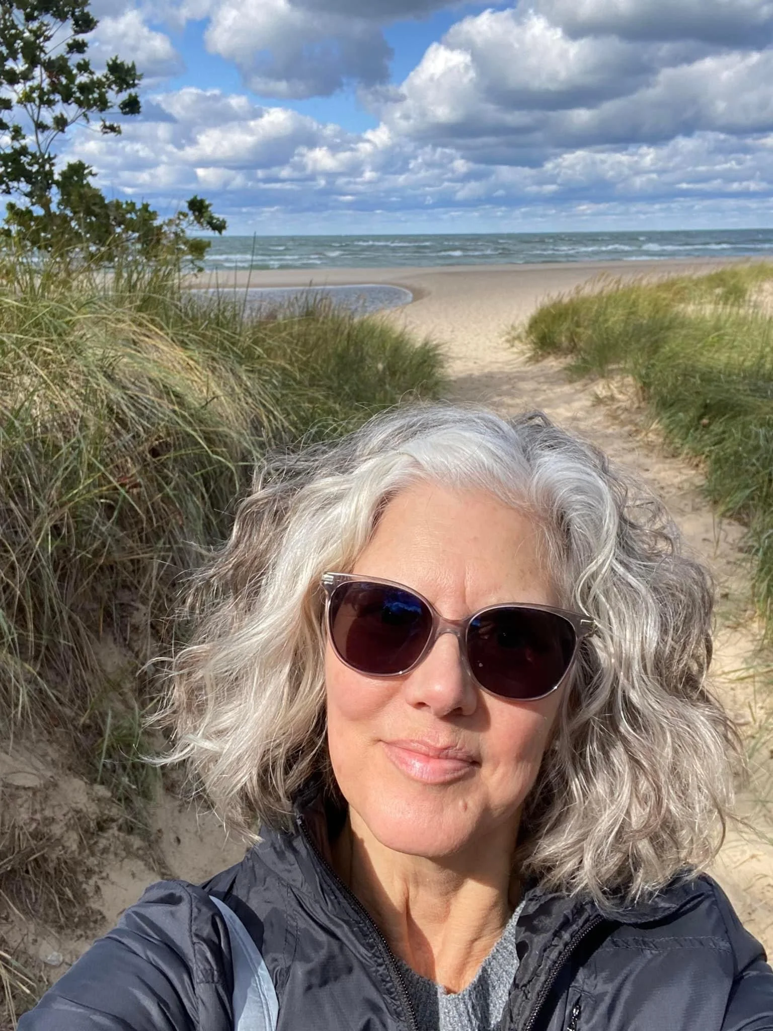 A woman with gray, curly hair wearing sunglasses and a black jacket taking a selfie at the beach, with sand dunes, grassy plants, and the ocean under a cloudy sky in the background.