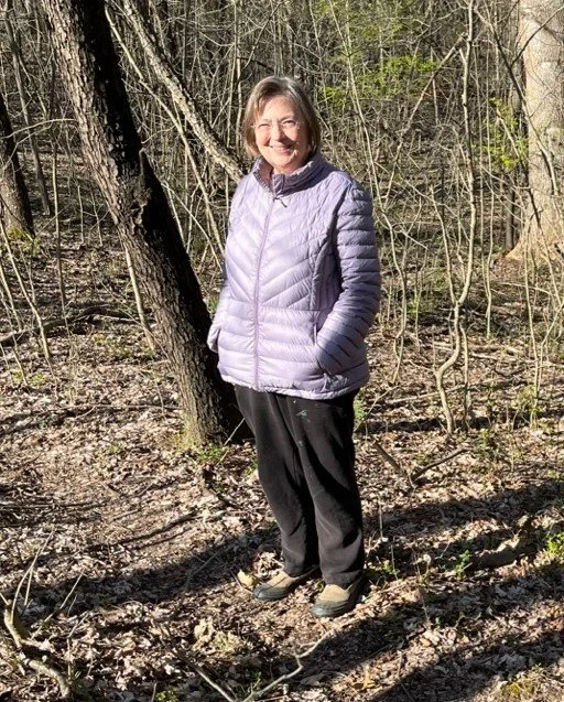 A smiling woman stands in a wooded area wearing a purple quilted jacket, black pants, and hiking shoes.
