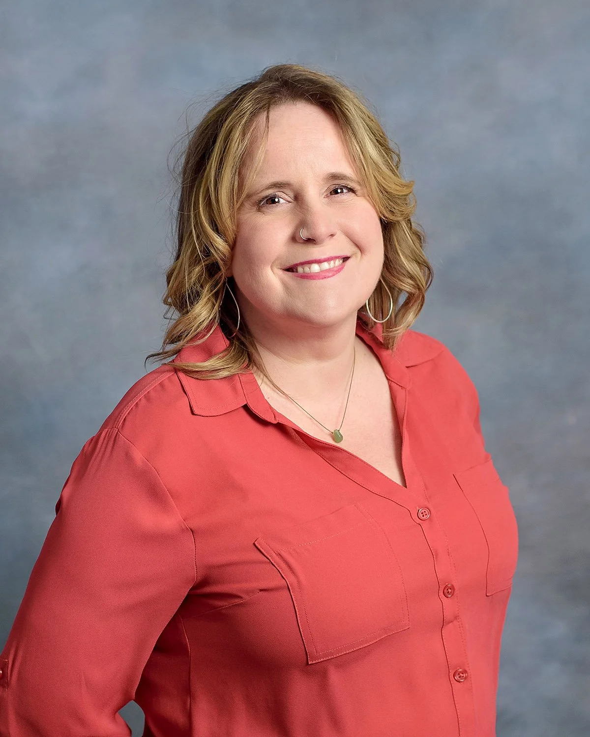 A woman with shoulder-length blonde hair smiling, wearing a coral button-up shirt and a necklace, standing in front of a gray background.