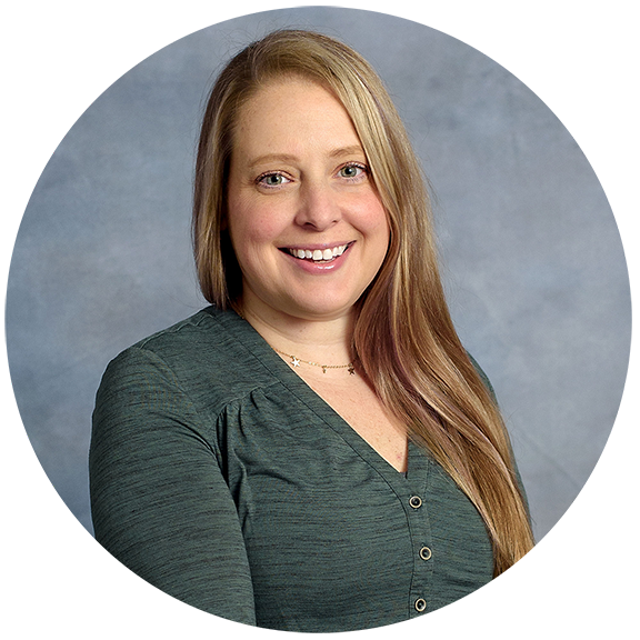 A woman with long, light brown hair smiling, wearing a dark green top against a gray background.