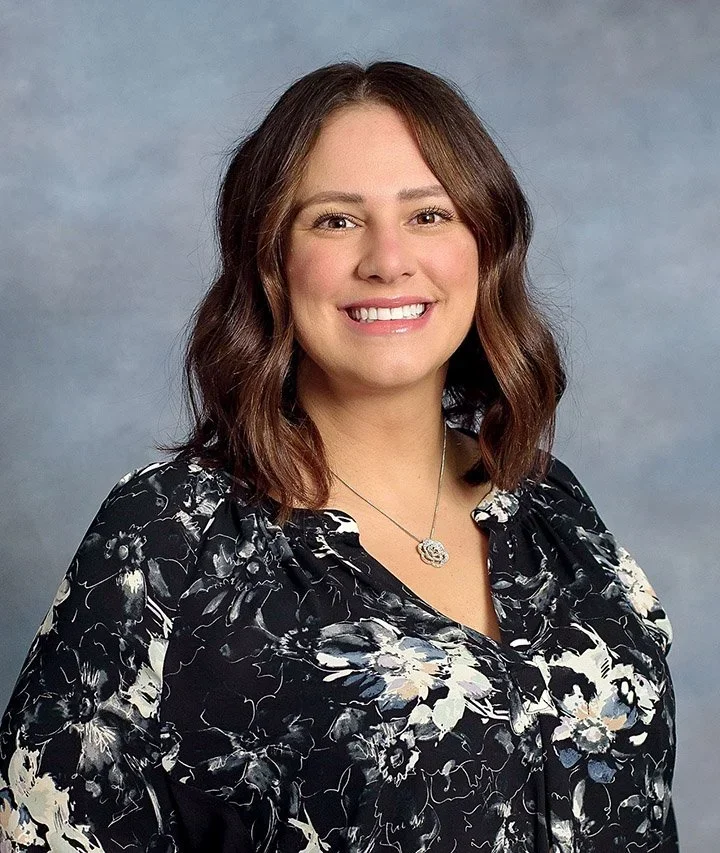 Professional headshot of a woman with shoulder-length brown hair, smiling, wearing a black floral blouse and a silver necklace, against a gray background.