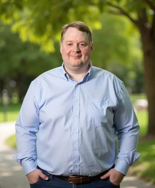 Man with short hair wearing a light blue button-up shirt standing outdoors in a park with green trees and sunlight in the background.