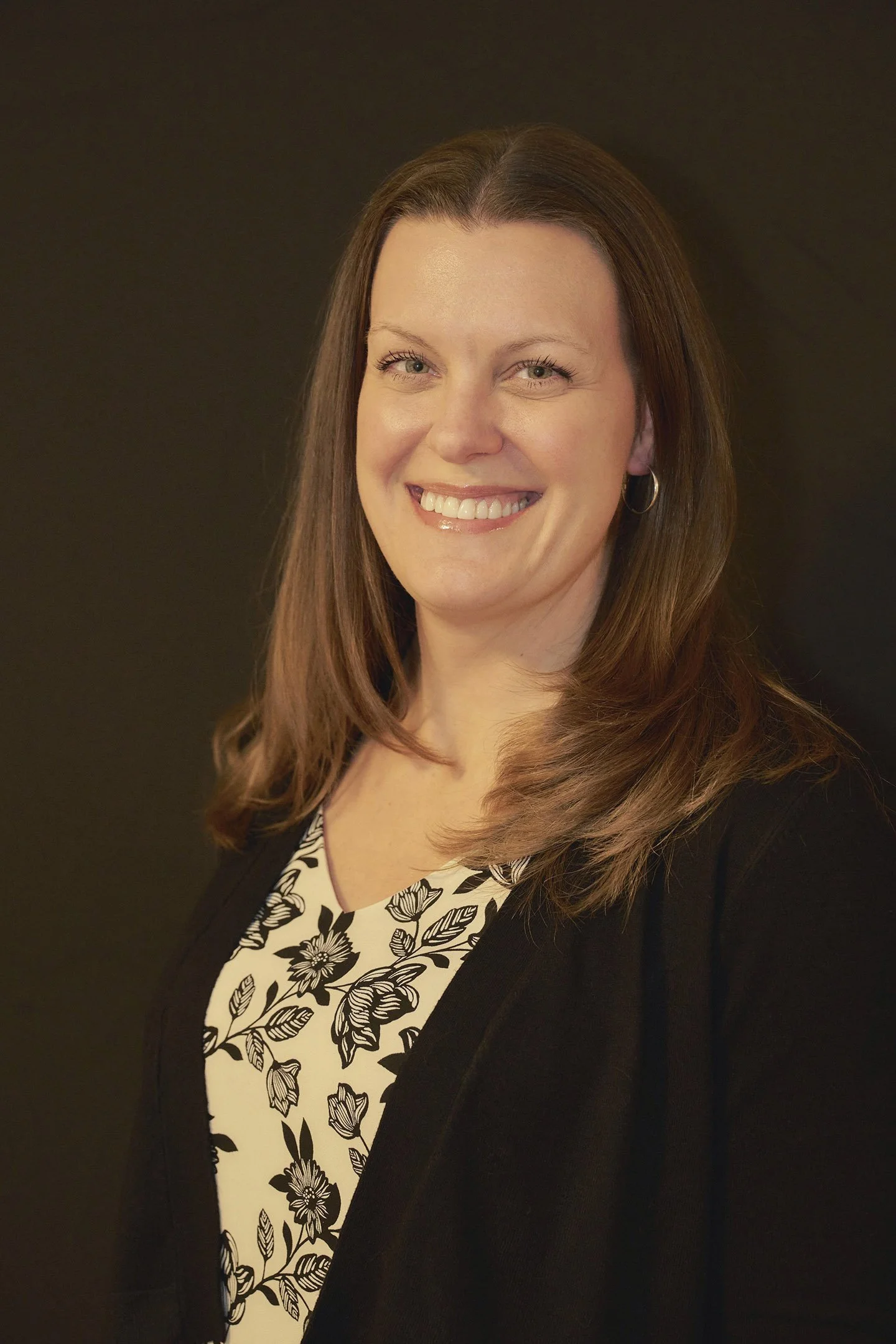 A smiling woman with shoulder-length brown hair, wearing a black blazer over a floral-patterned blouse, standing against a dark background.