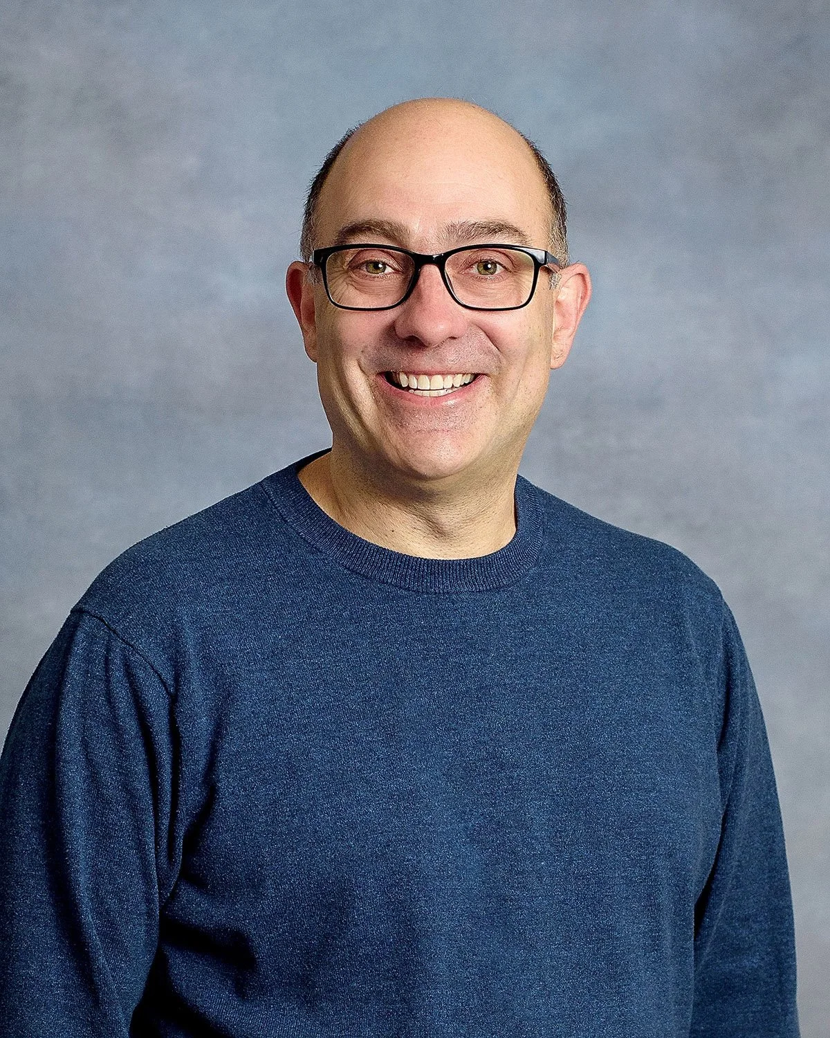 A smiling man with glasses, short hair, and a blue sweater standing in front of a grey background.