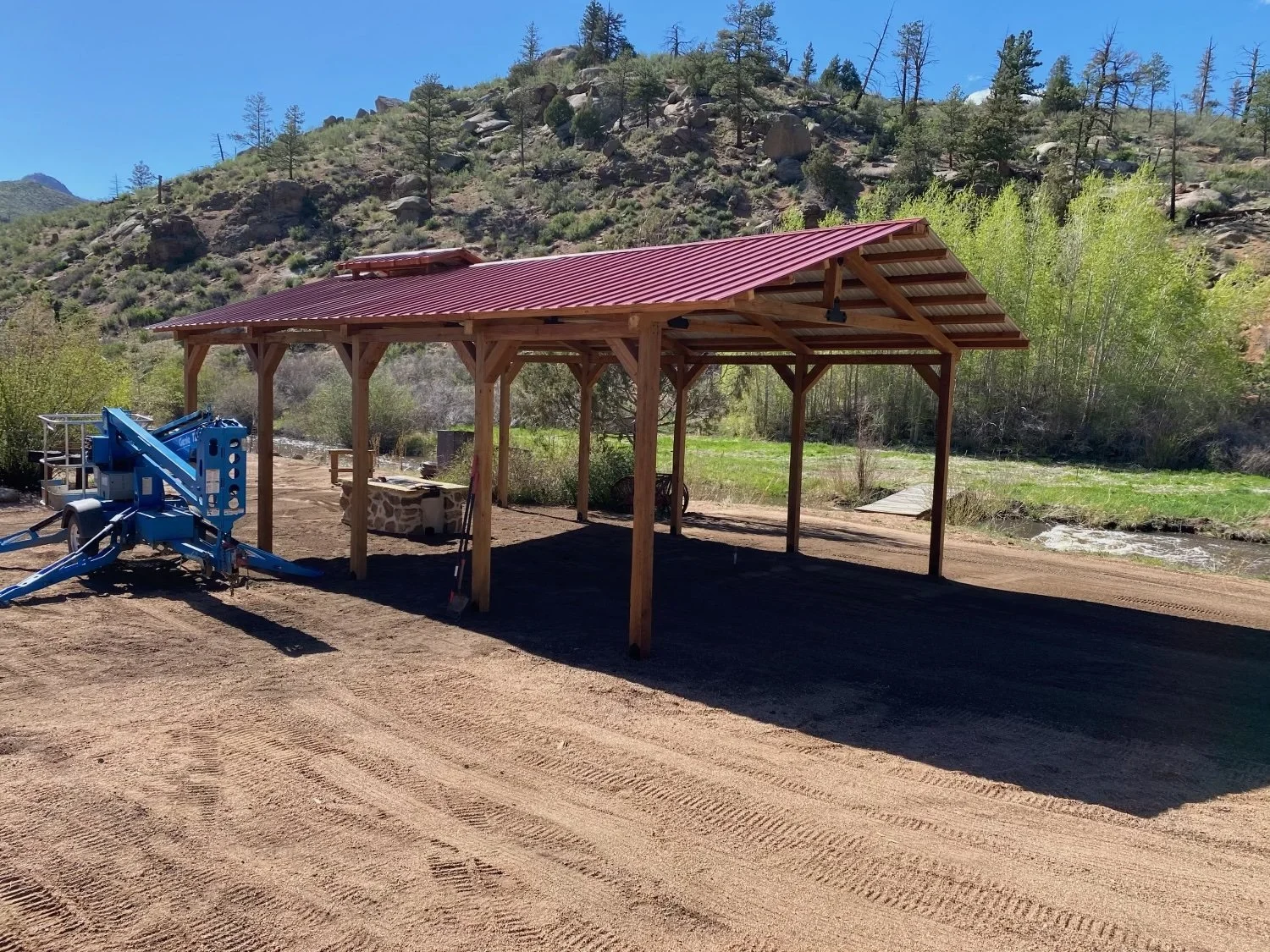 A newly constructed wooden pavilion with a red metal roof in a rural outdoor setting, with a mountain and trees in the background, and dirt ground in the foreground.