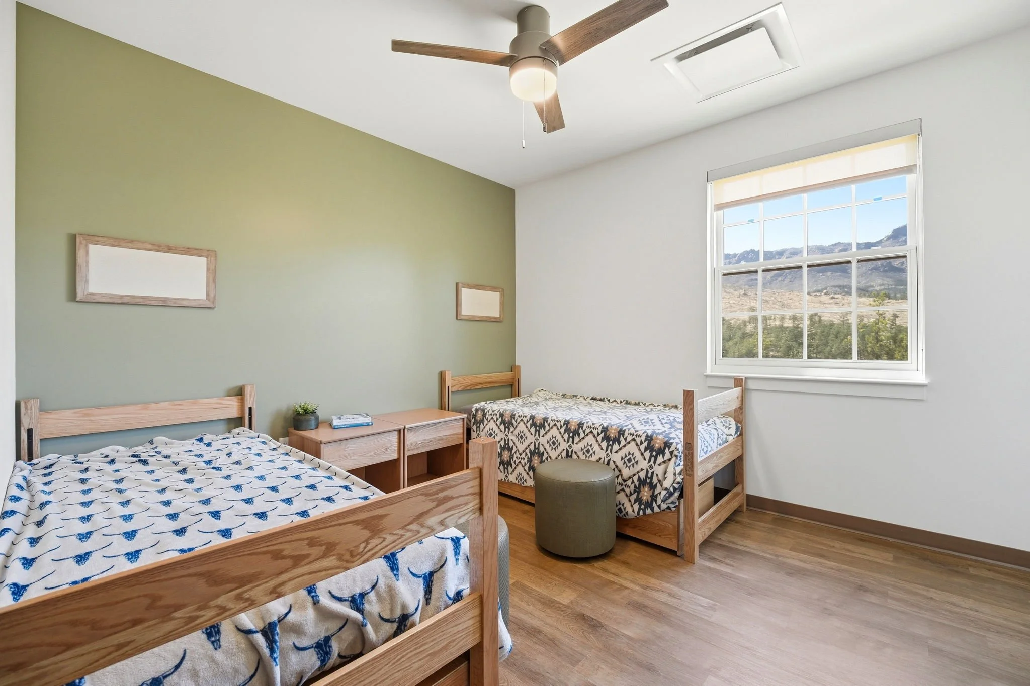 A bright bedroom with two wooden beds, one with a patterned comforter and the other with a blue and white design. A wooden nightstand between the beds has a small plant and a few books. A large window shows a view of mountains and blue sky, with beig