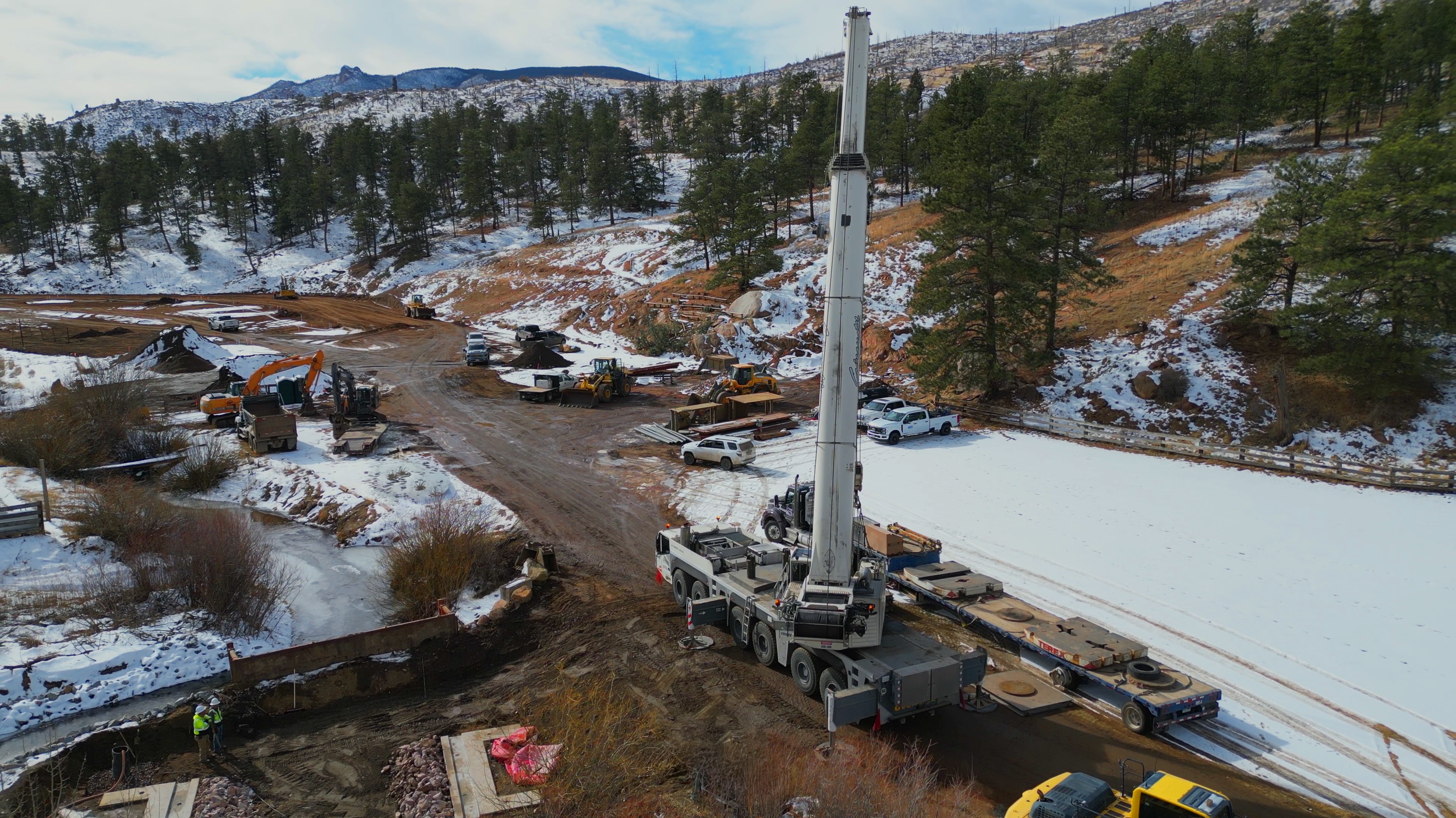 Construction site in a mountainous, snowy area with various heavy machinery, trucks, and workers