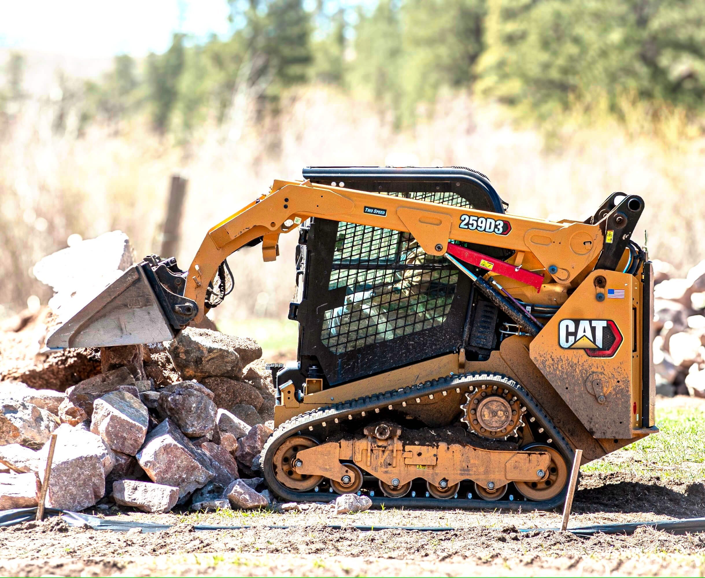 A yellow Caterpillar mini excavator working on a rocky outdoor site, with a blurred background of trees.