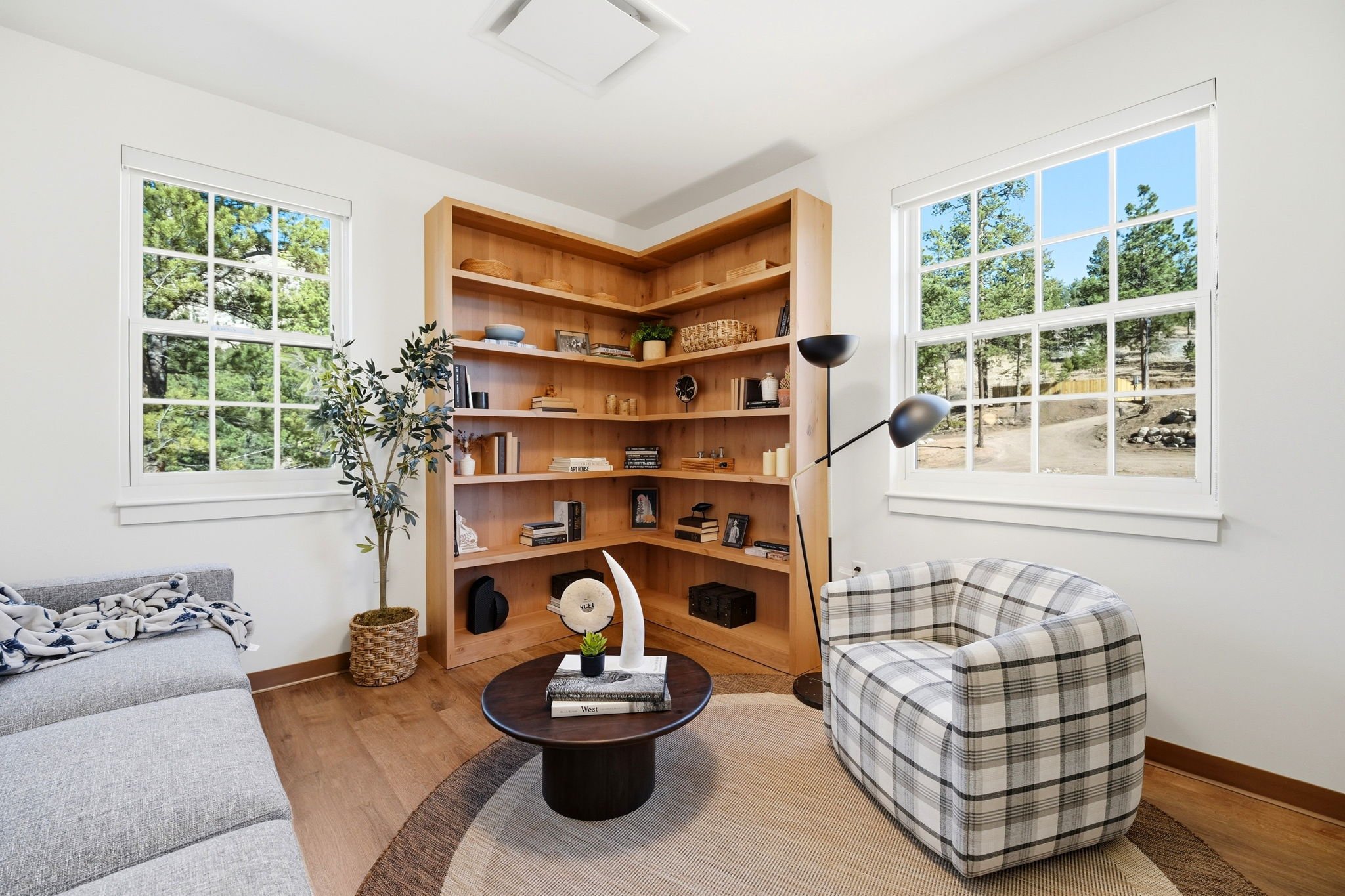 Living room with two large white-framed windows showing trees outside, a wooden bookshelf filled with books and decorative items, a black floor lamp, a gray sofa with a blanket, a plaid armchair, and a round coffee table with books and a small plant.