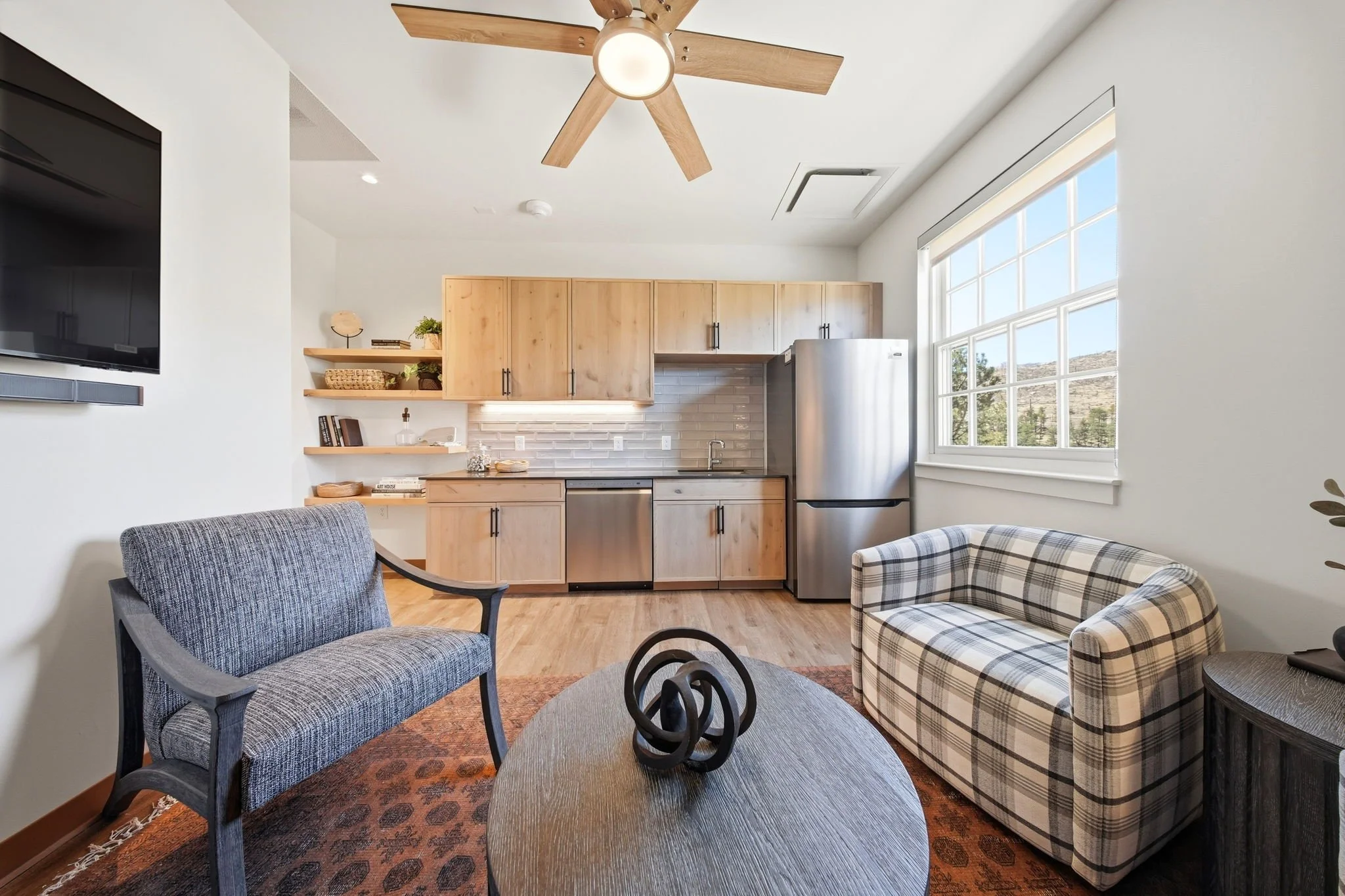 An open-concept living room and kitchen with natural light, modern furniture, wooden cabinetry, a ceiling fan, and a large window showing an outdoor landscape.