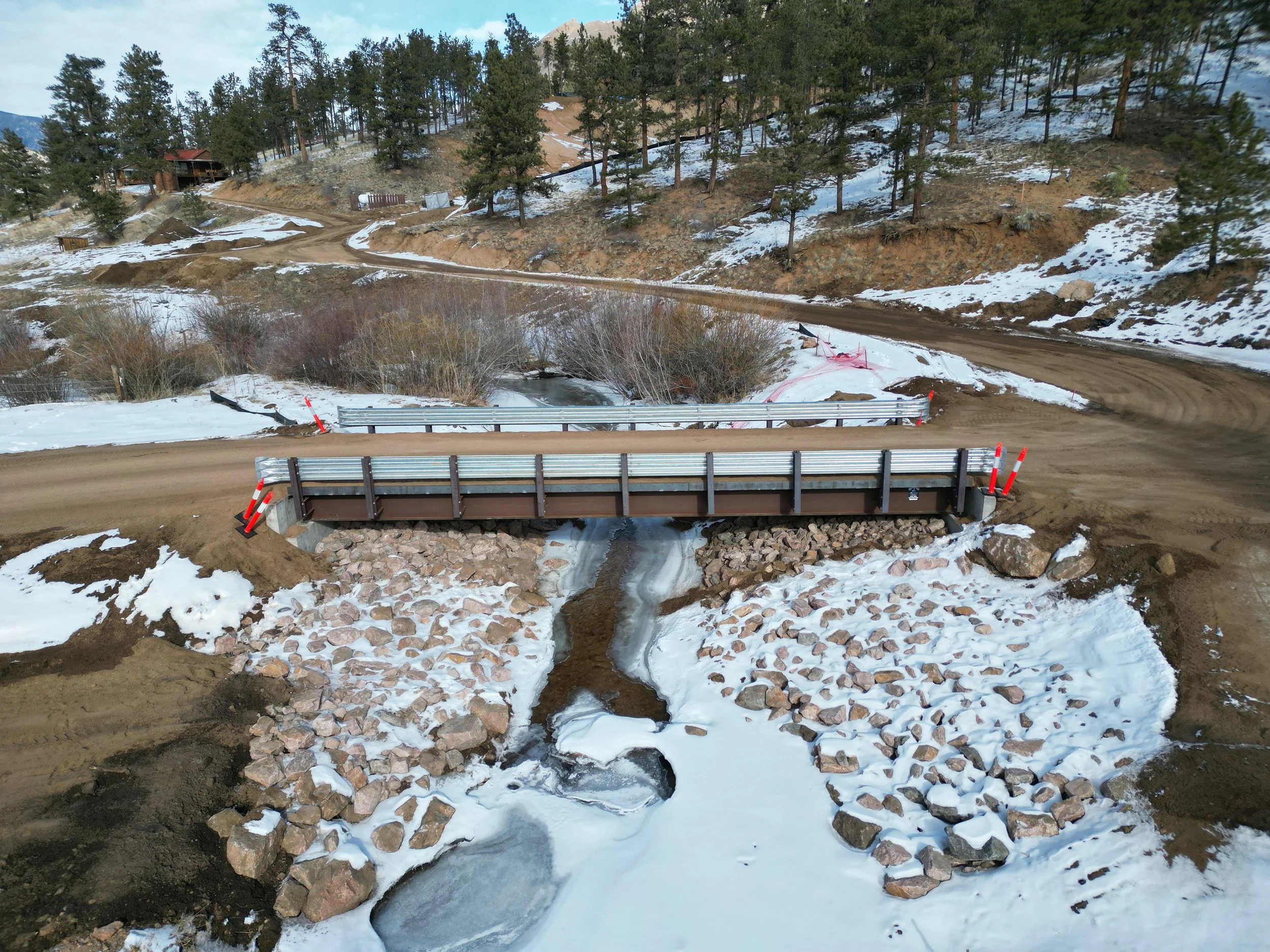 A bridge over a small stream on a dirt road with snow and dirt on the ground. There are trees on a hillside in the background and a few houses on the hillside.