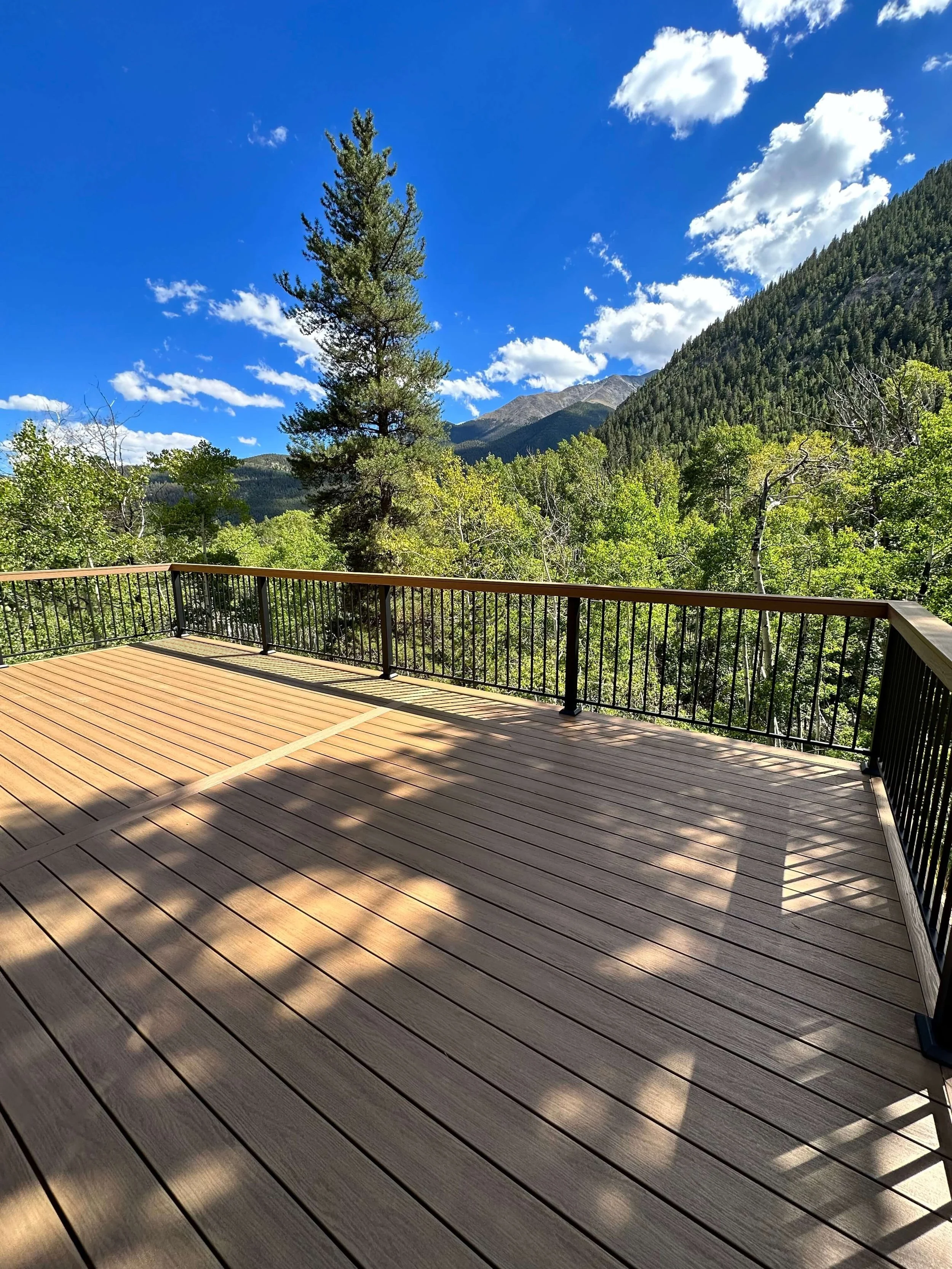 A wooden deck with a black metal railing overlooking a lush green forest with trees, mountains, and a clear blue sky with fluffy white clouds.