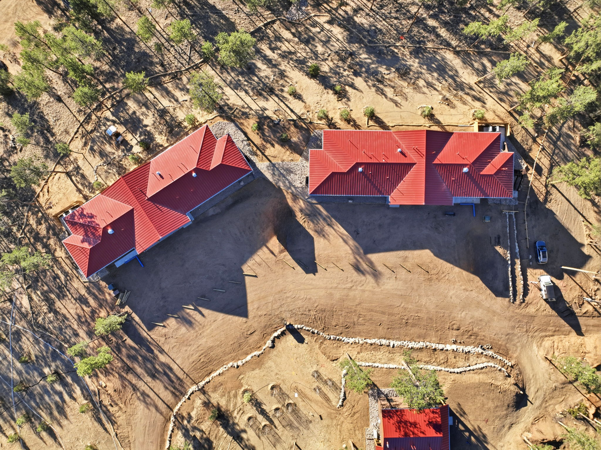 An aerial view of a house with a red metal roof, surrounded by a dirt landscape and sparse trees, with a gravel driveway and cars parked nearby.