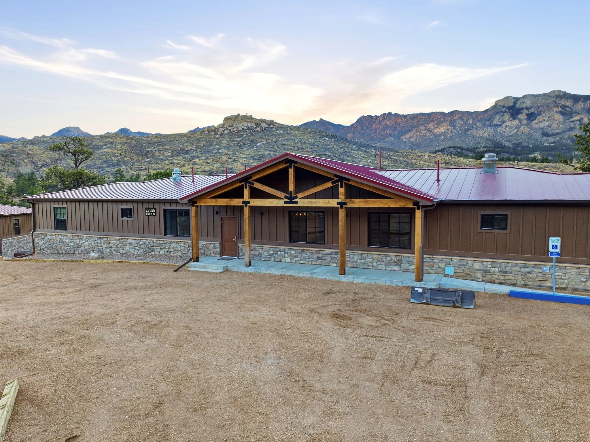 A newly constructed modern house with a red metal roof and wooden beams at the front, set against a mountainous landscape with green shrubbery, under a partly cloudy sky.
