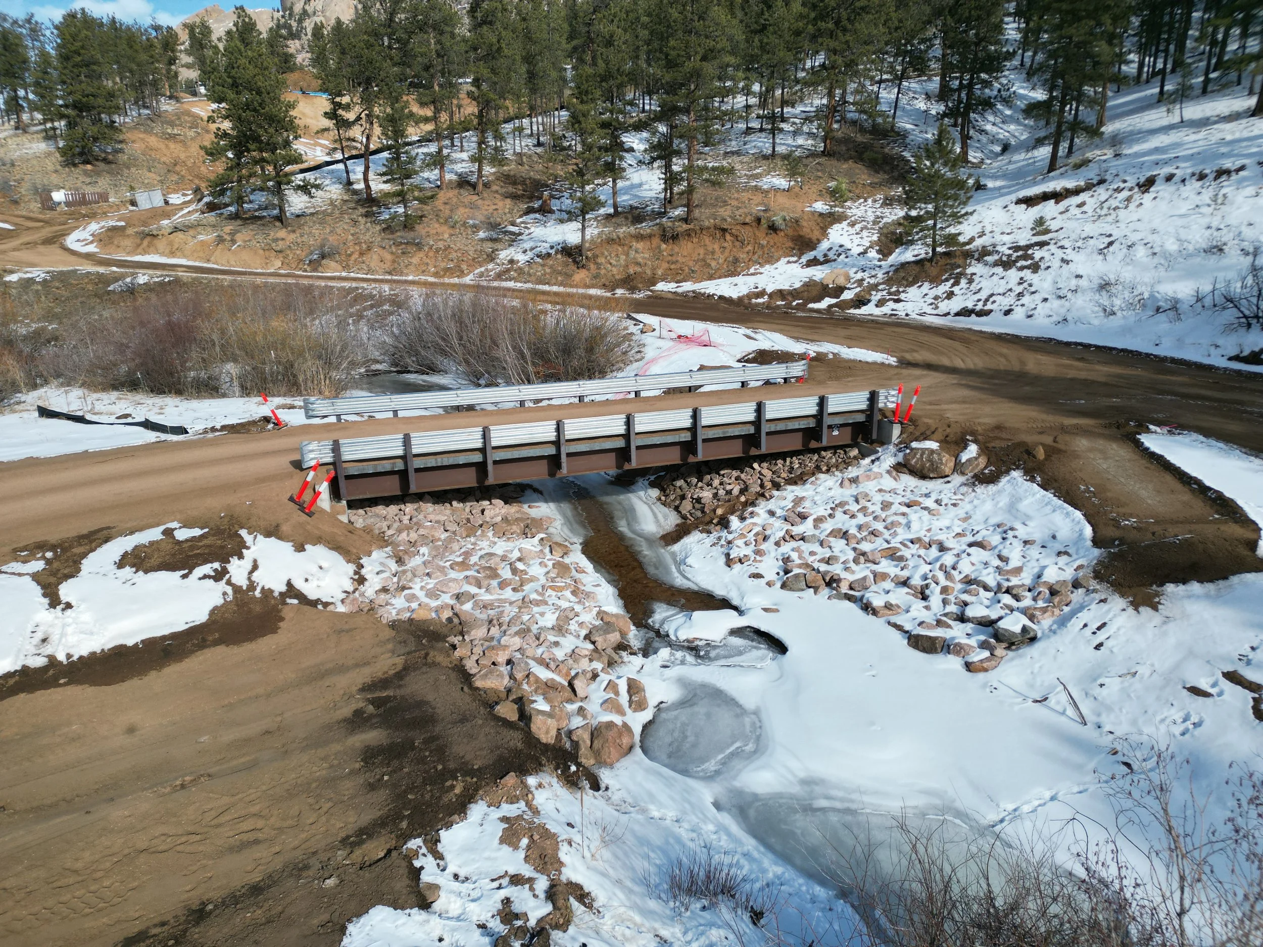 A dirt road in a mountainous area with snow on the ground and trees on either side, and a small creek flowing underneath a new bridge.