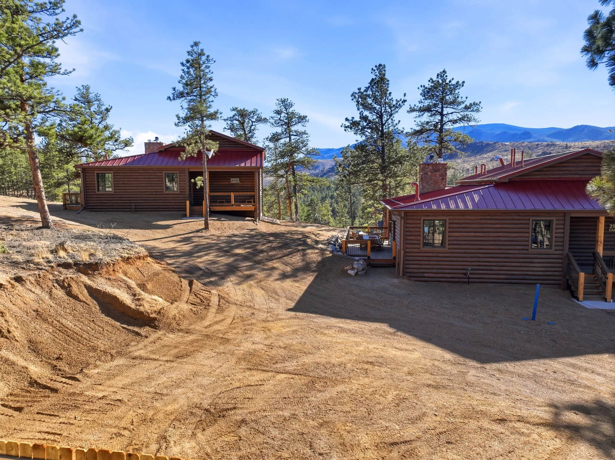 Snapshot of a residential area with two log cabin-style houses featuring red roofs set among tall pine trees, with mountain range visible in the background on a clear, blue sky day.