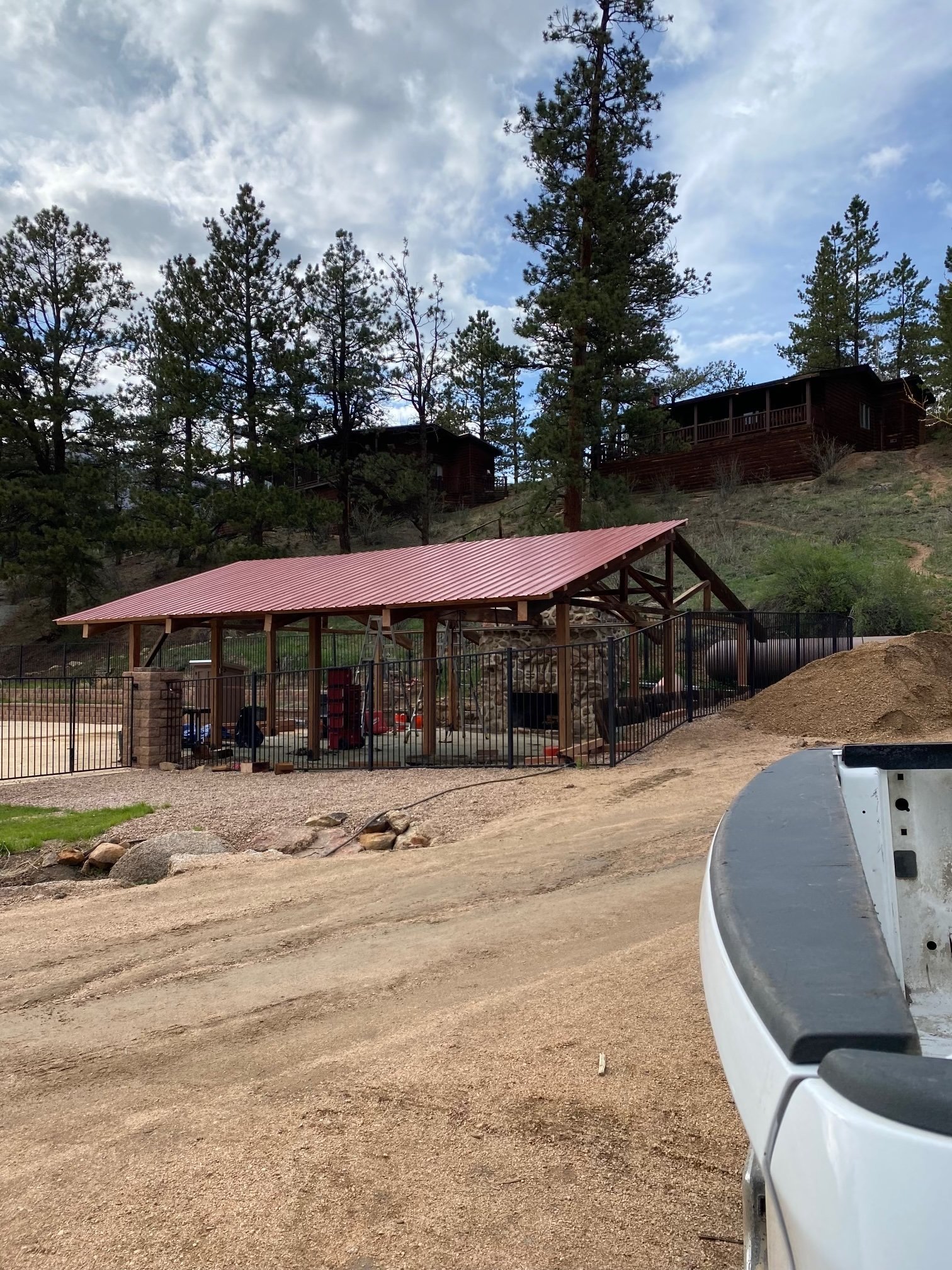 Parked vehicle in the foreground with a construction site behind, showing a dirt pathway, a black metal fence, a red-roofed shelter, and multiple cabins on a hillside among tall pine trees with a partly cloudy sky.