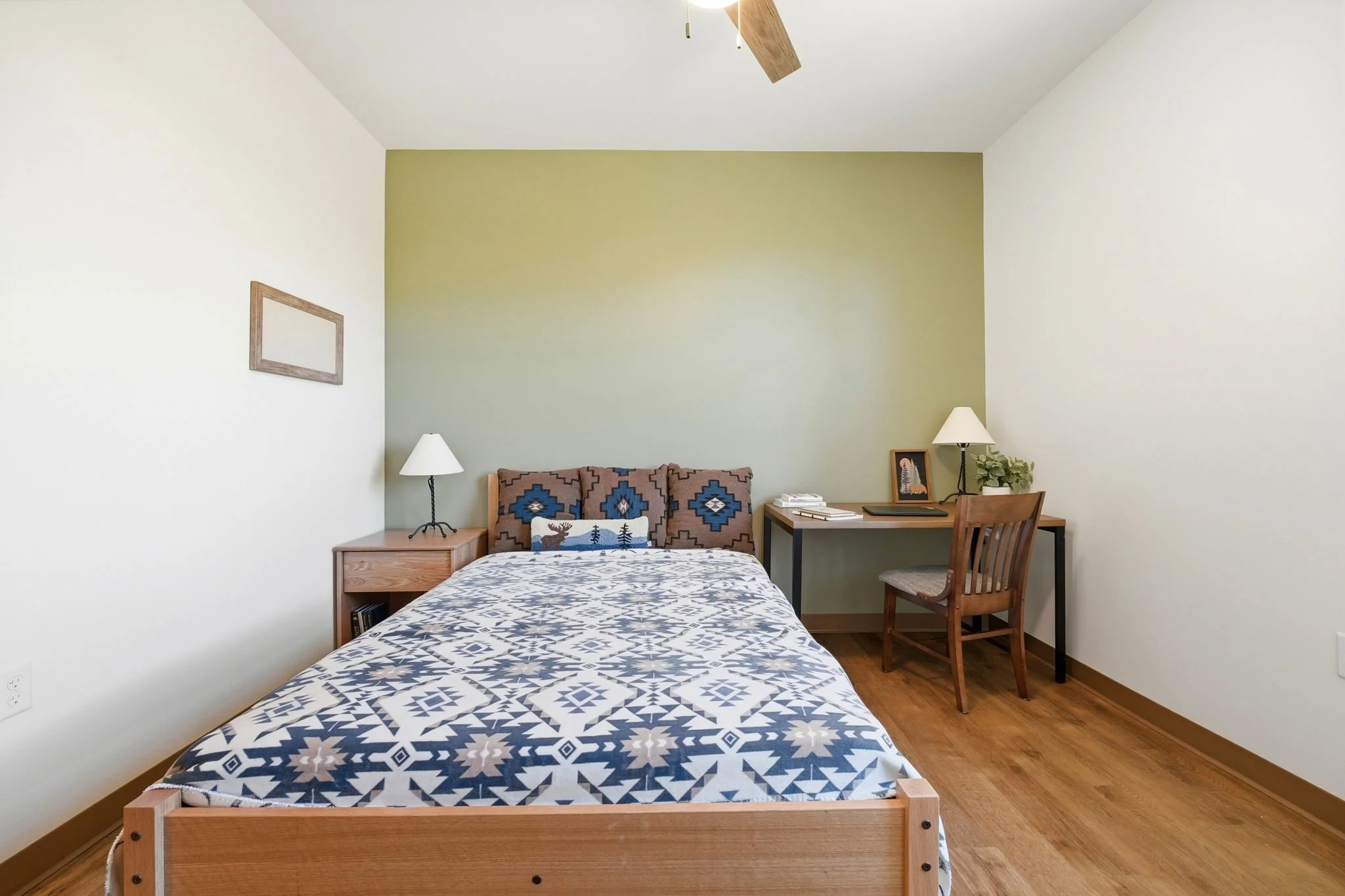 A bedroom with a wooden bed, patterned bedding, two nightstands with lamps, a small desk with books, a chair, framed pictures, and a ceiling fan. The wall behind the bed is painted green, and the other walls are white.