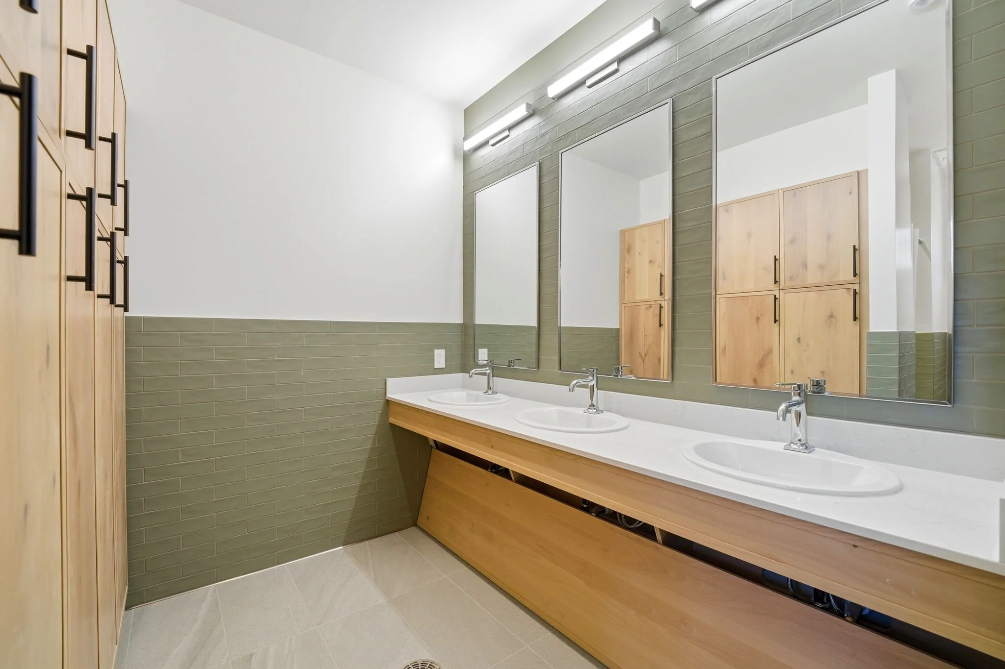 Modern bathroom with double sinks, large mirrors, wooden cabinet, and green tile walls.