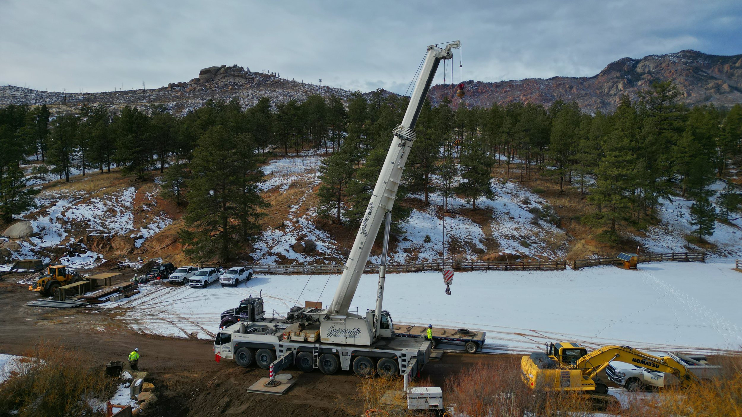 Construction workers using a crane on a snowy hillside during daytime, with parked cars and construction equipment nearby, and a mountainous landscape in the background.