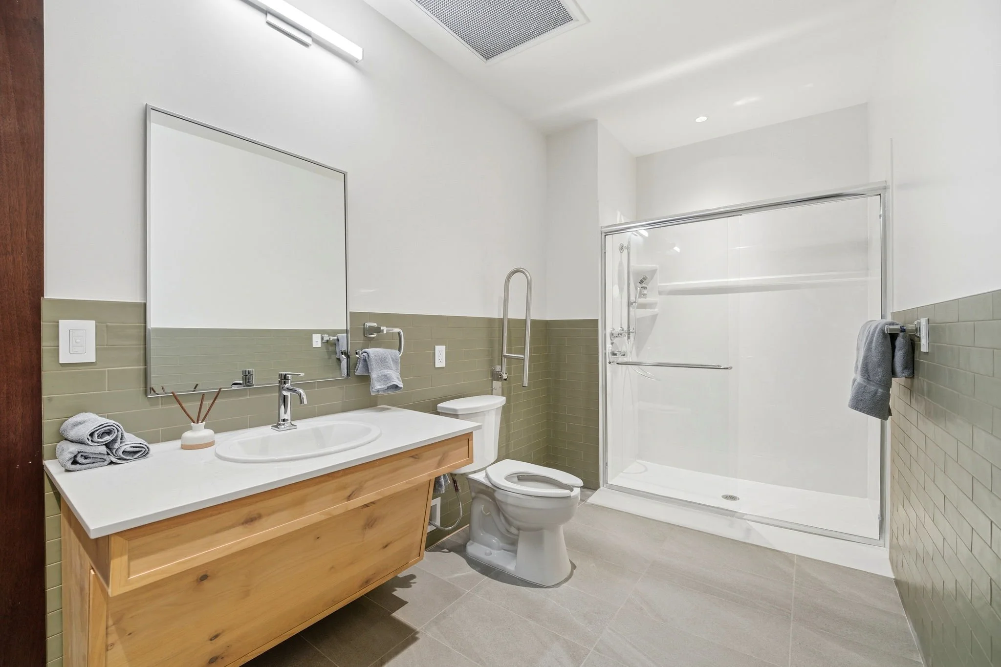 Modern bathroom with a wooden vanity, white countertop, round sink, mirror, gray towels, a shower with a glass door, tiled wall, and ceiling light.