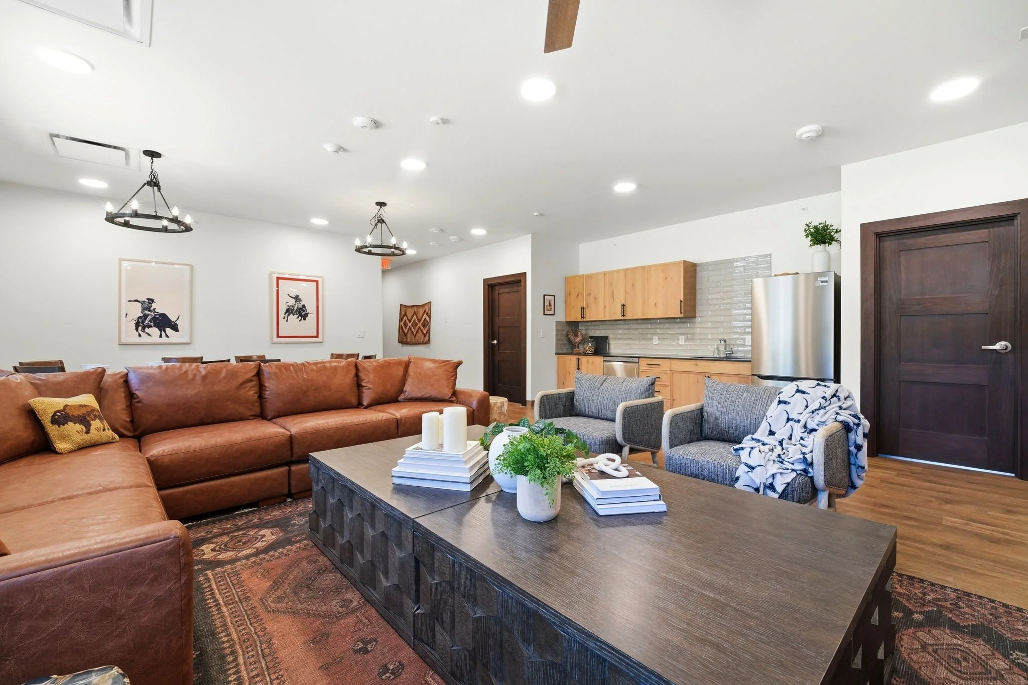 Living room featuring a large brown leather sectional sofa, a wooden coffee table with decorative objects and books, and a cozy seating area with two gray armchairs and a blanket. The room has white walls with framed artwork, wooden cabinets in the k