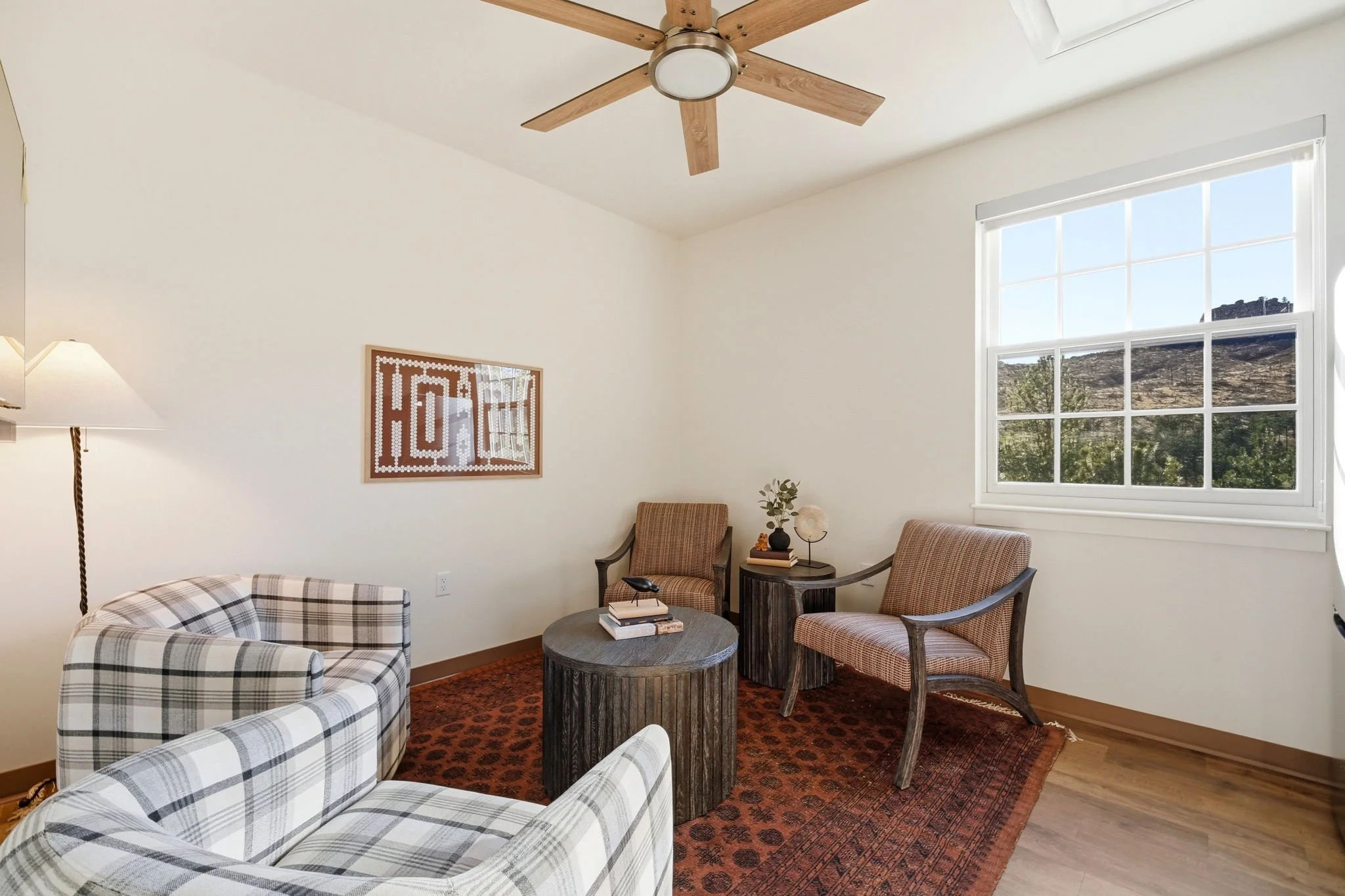 Living room with white walls, large window with scenic view, ceiling fan, framed artwork, plaid armchair, two wicker chairs, round wooden coffee table, small side table with plants and decor, standing lamp, area rug.