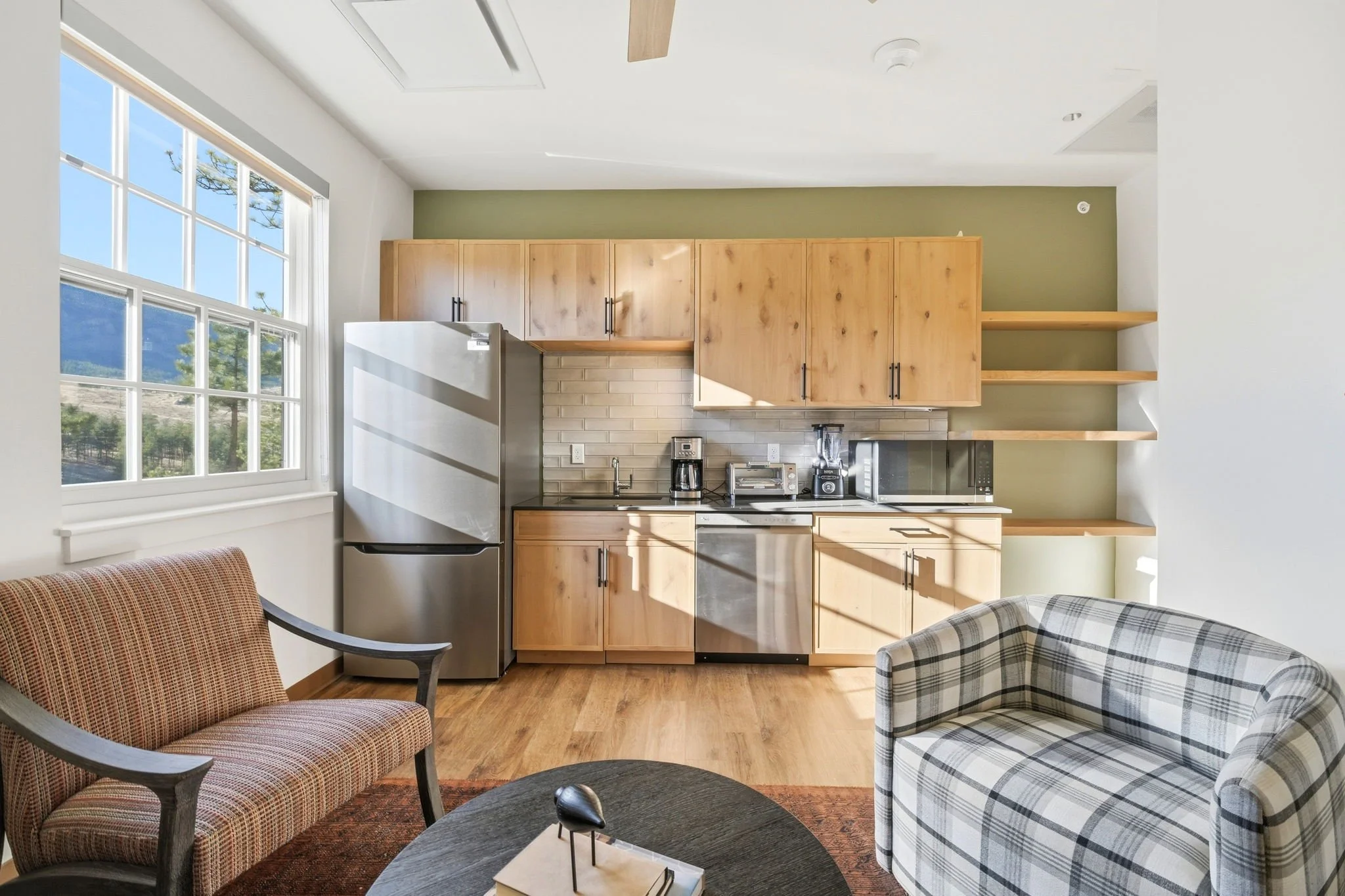 Living room with large window, wooden kitchen cabinets, stainless steel refrigerator, countertop appliances, armchair, and plaid armchair.