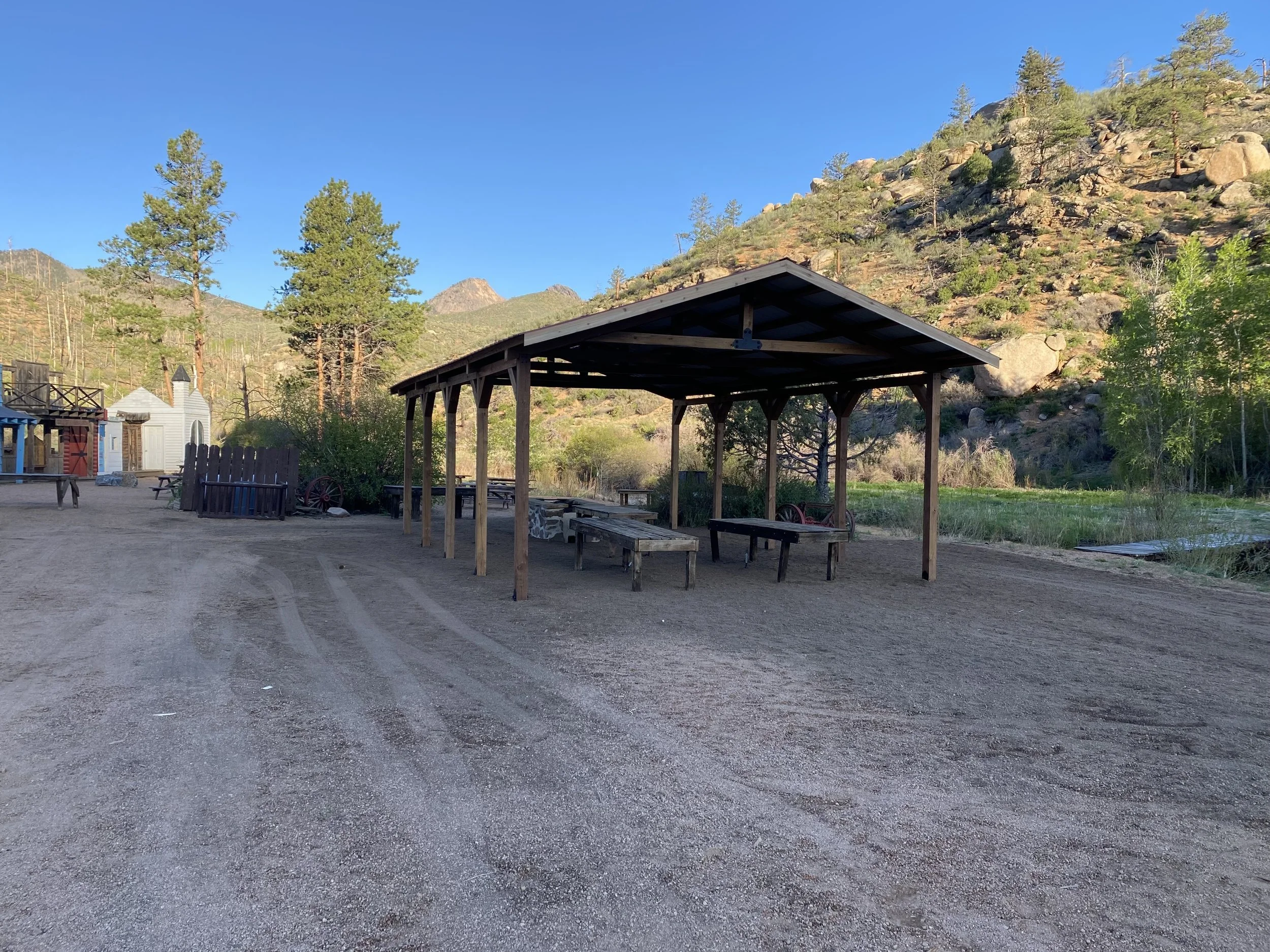 A rustic outdoor setting with a wooden pavilion and benches, surrounded by trees and a mountain hillside under a clear blue sky.