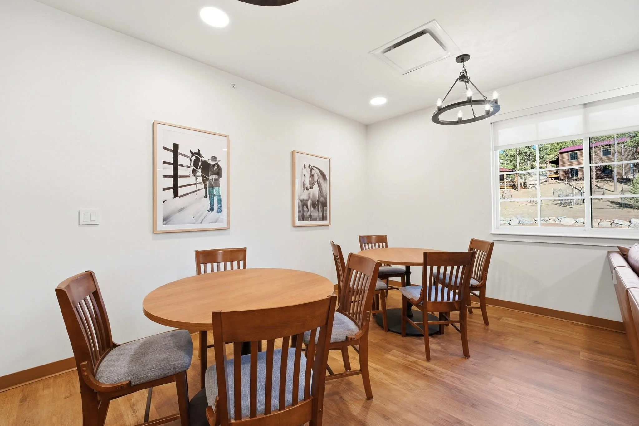 A dining area with a round wooden table and ten wooden chairs with gray cushions, a large window showing a view of houses and trees, white walls with two framed horse-themed pictures, and a modern chandelier with candles hanging from the ceiling.