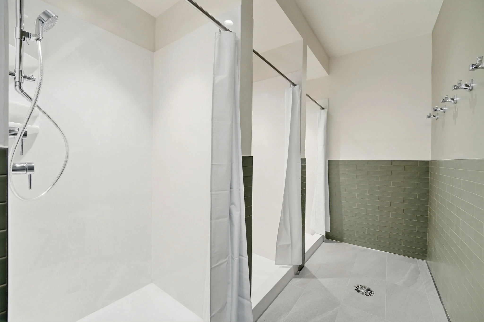 A modern shower area with white walls, a handheld showerhead, and a shower curtain. The floor has light gray tiles, and there is a drain on the floor. To the right, the wall has a section with greenish tiles.