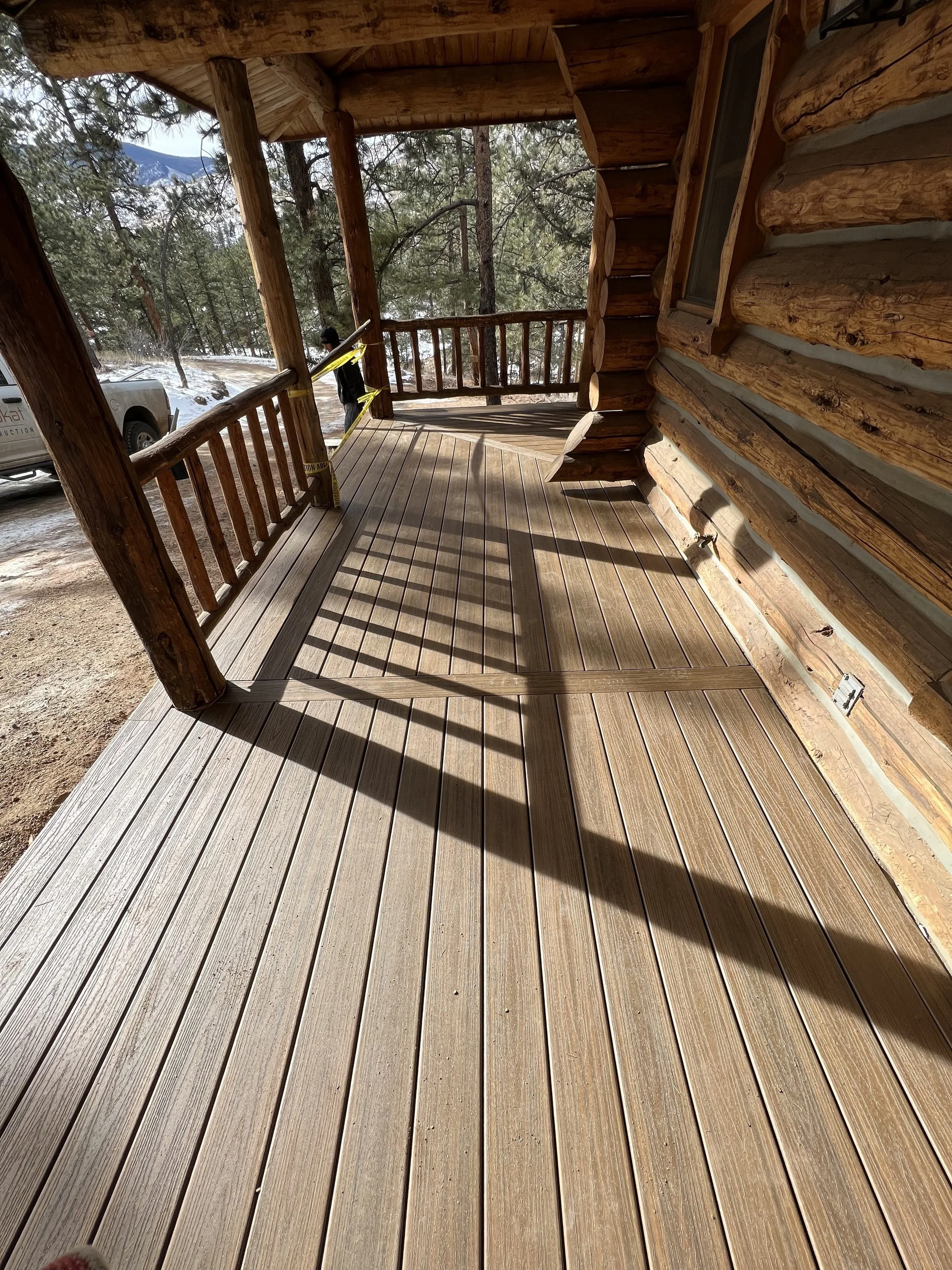 A wooden porch attached to a log cabin with wooden railing, overlooking a forested area with snow on the ground and mountains in the distance.