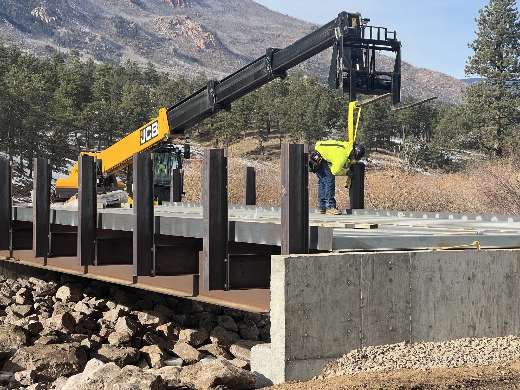 Construction site with a yellow JCB crane lifting a black forklift, workers in safety gear, and rocky terrain with mountain and trees in the background.