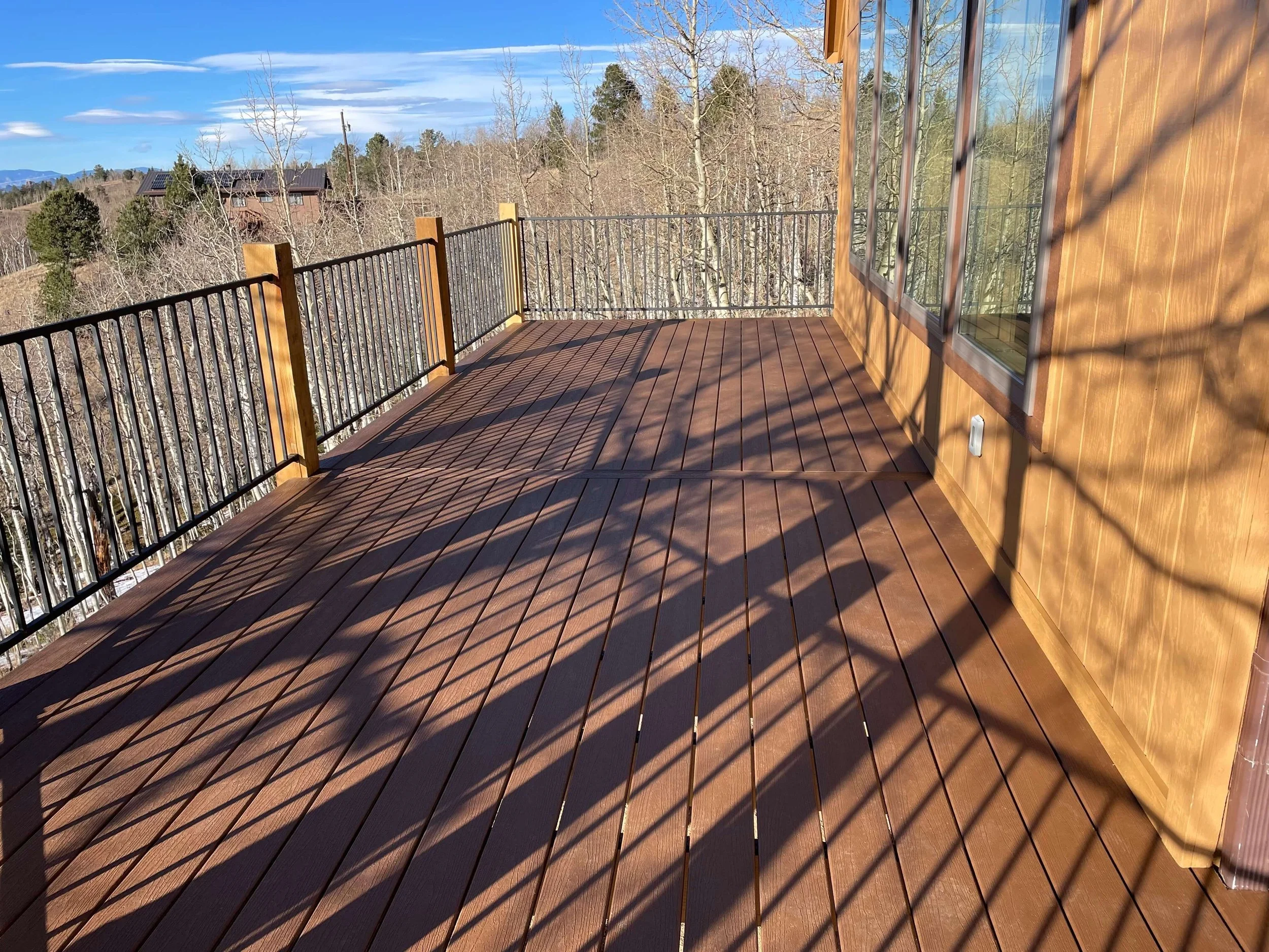 A wooden balcony with black metal railing surrounded by leafless trees and distant houses with mountains in the background under a partly cloudy sky.