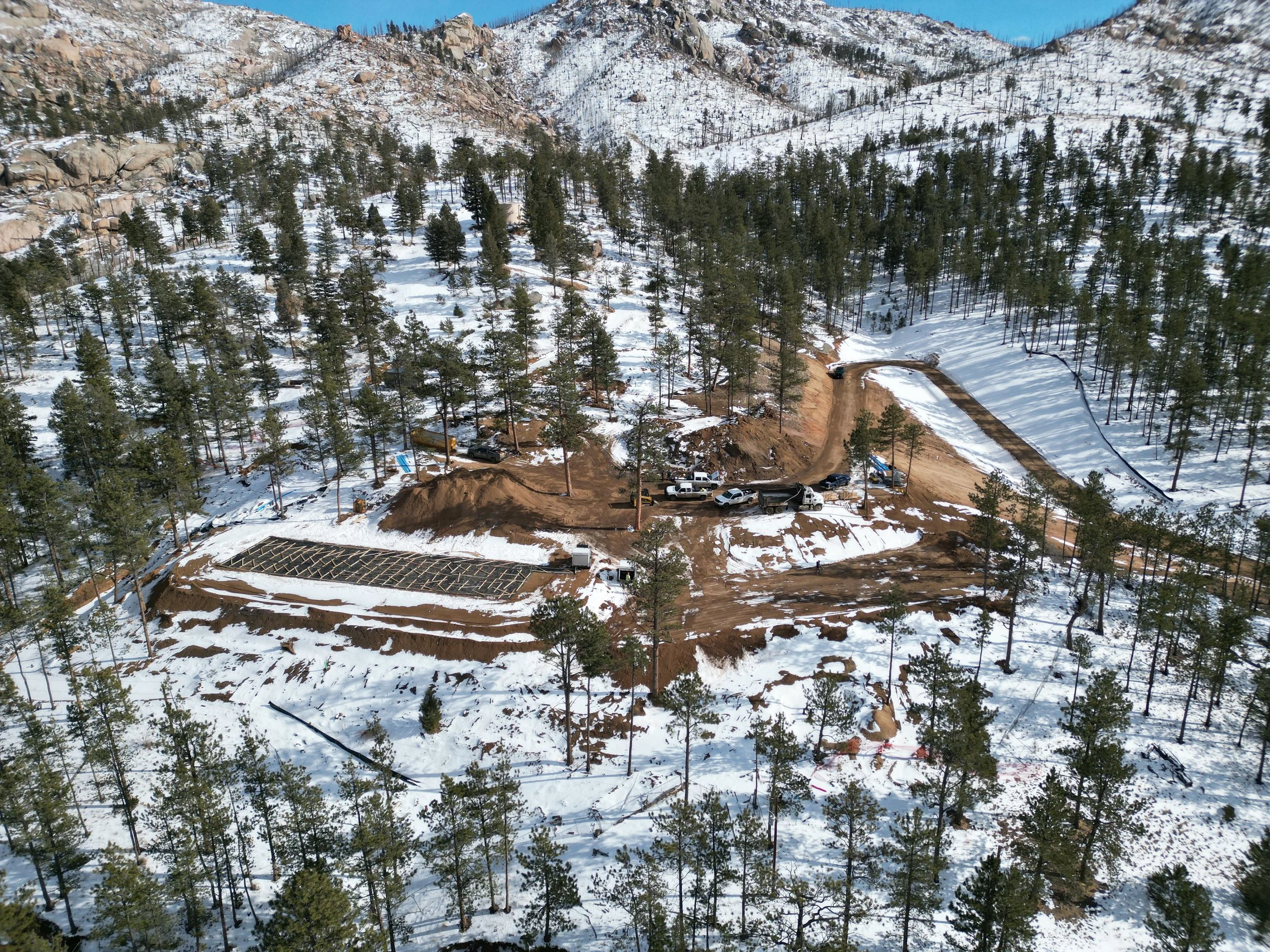A snow-covered mountainous landscape with many tall trees and a clear blue sky. There is a dirt road winding through the snow, with some parked cars and construction equipment at the bottom of the image.