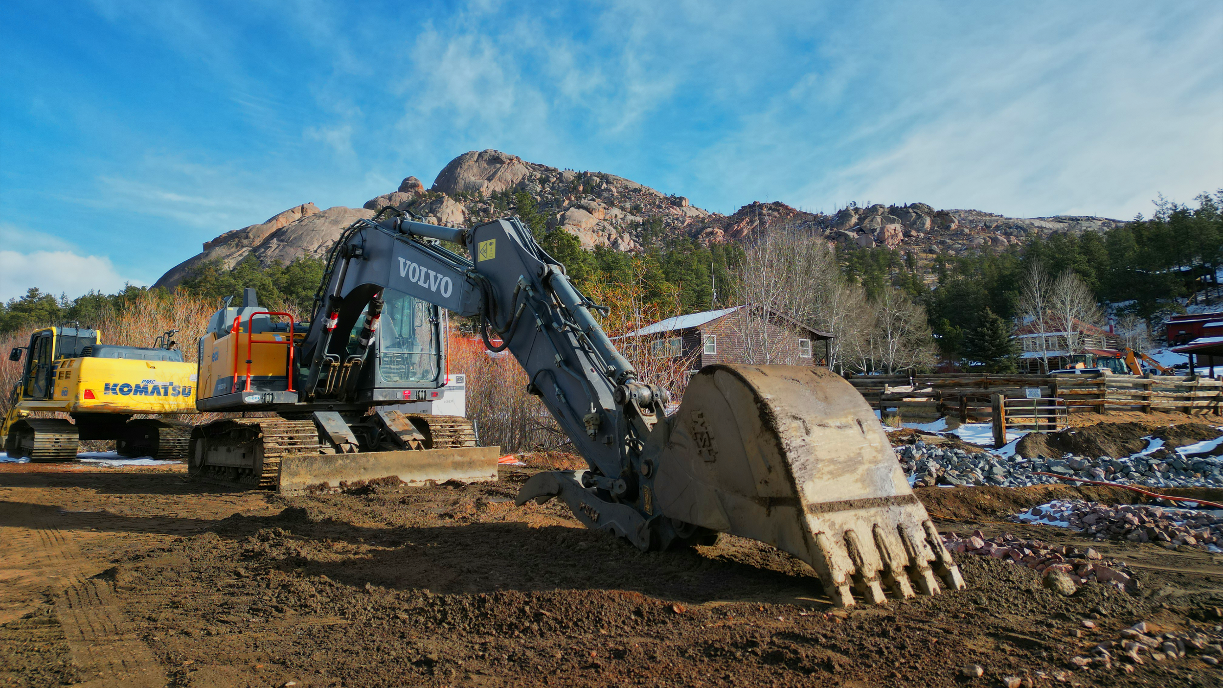 Construction site with a large excavator labeled Volvo and a smaller Komatsu excavator, dirt and rocks on the ground, trees, and houses with a mountain in the background under a blue sky.